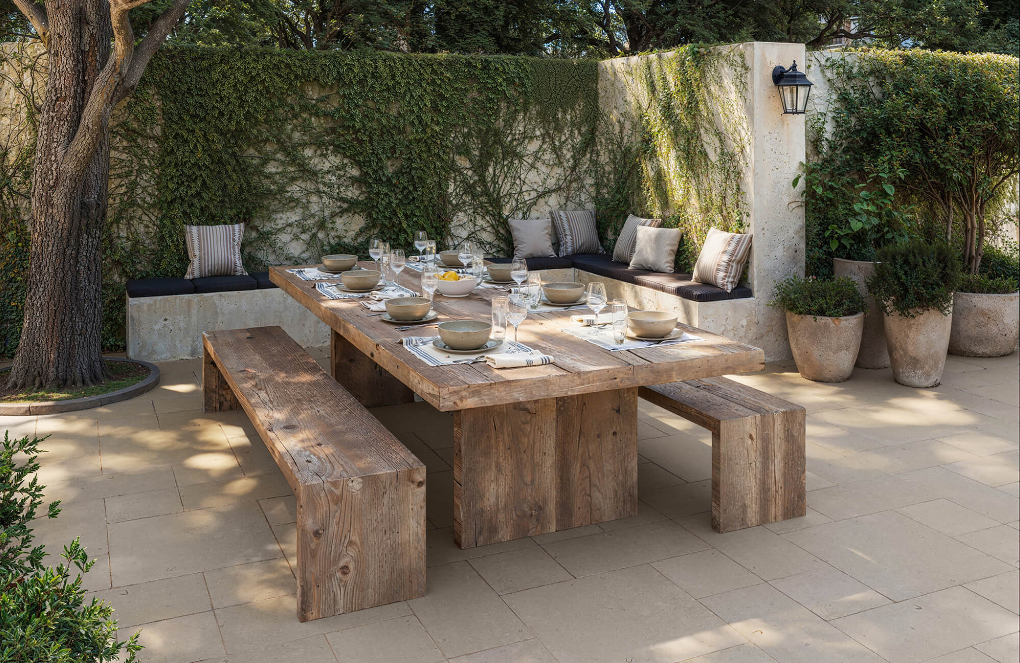 Rustic outdoor dining setup with a long wooden table, matching benches, built-in corner seating, place settings, glassware, and garden walls covered in vines.