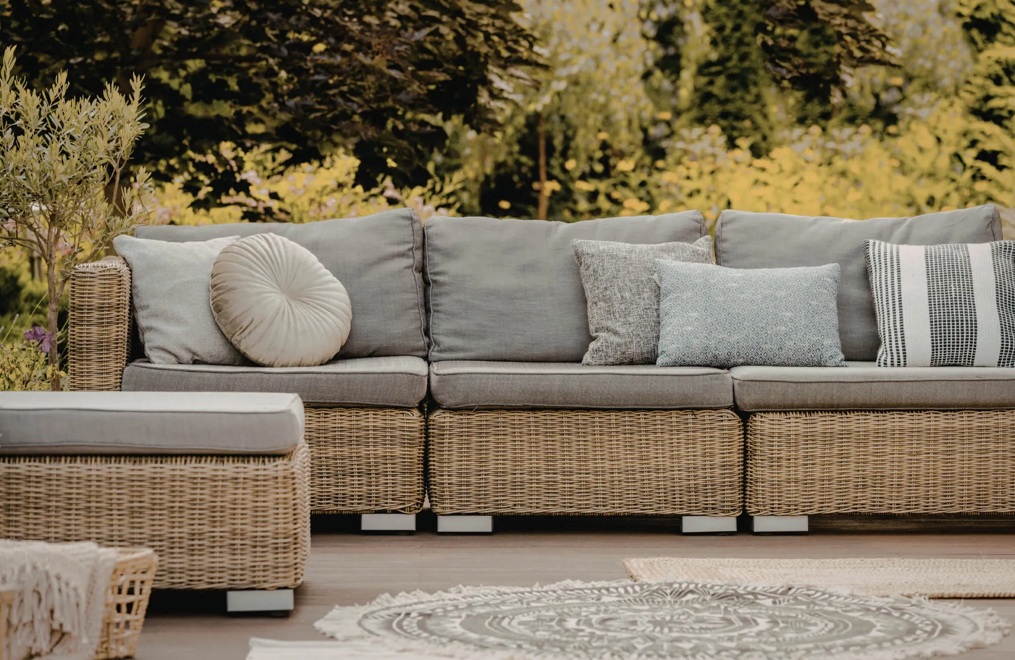 Outdoor wicker sectional sofa with mixed decorative pillows in neutral tones on a sunlit patio surrounded by greenery.