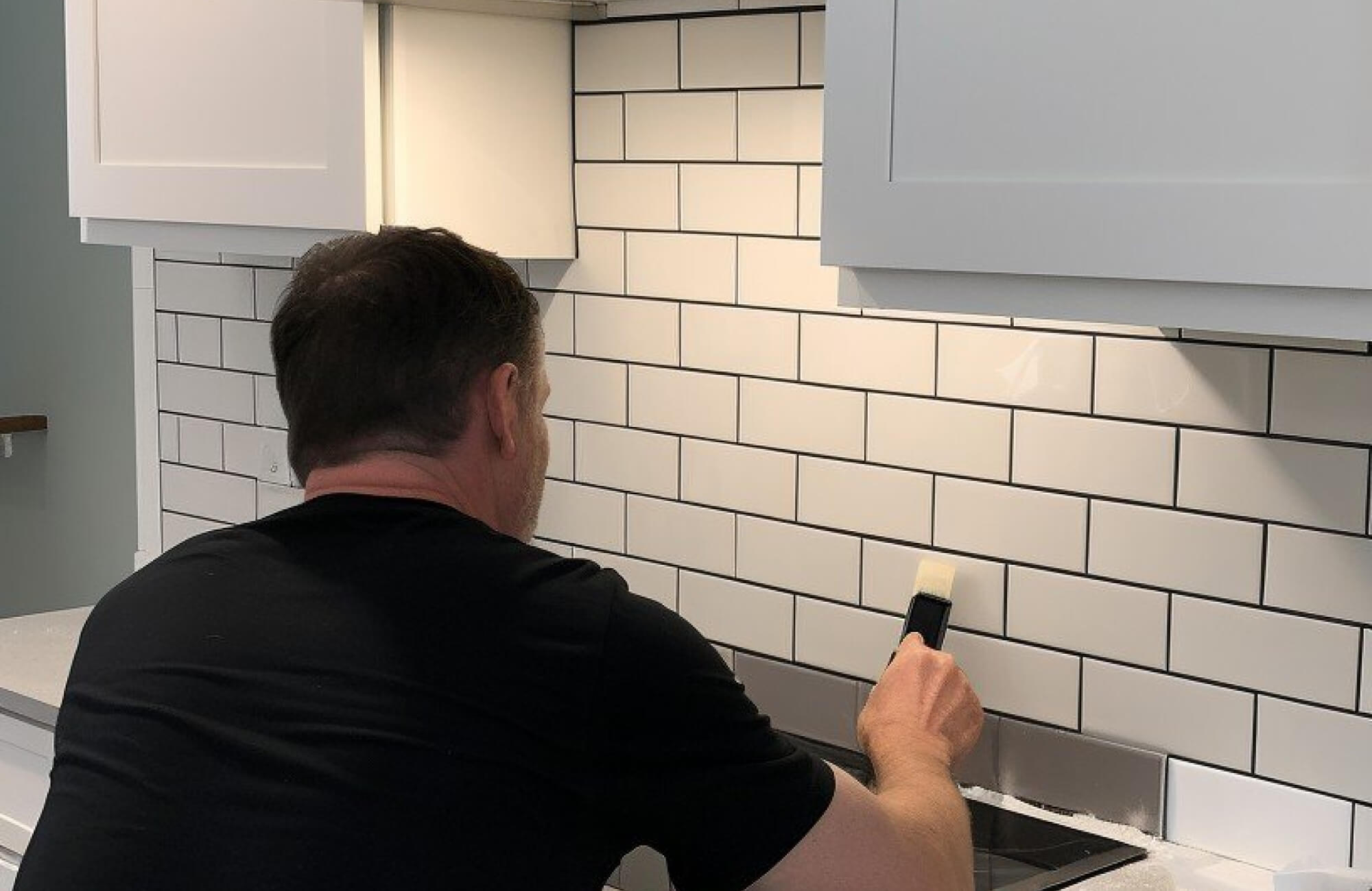 null An installer working on a white subway tile backsplash with dark grout lines in a modern kitchen.