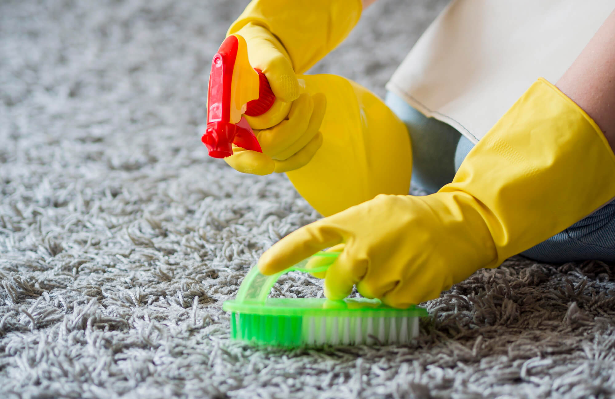 null A Person wearing yellow gloves is scrubbing a gray rug with a spray bottle and brush to remove stains effectively.