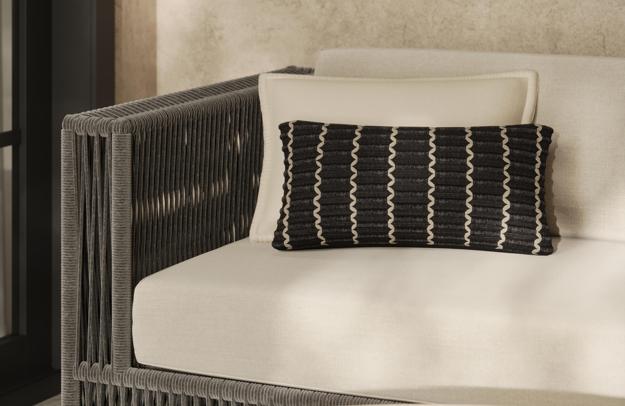 Black patterned lumbar pillow layered on a cream outdoor lounge chair with woven gray rope arm detailing.