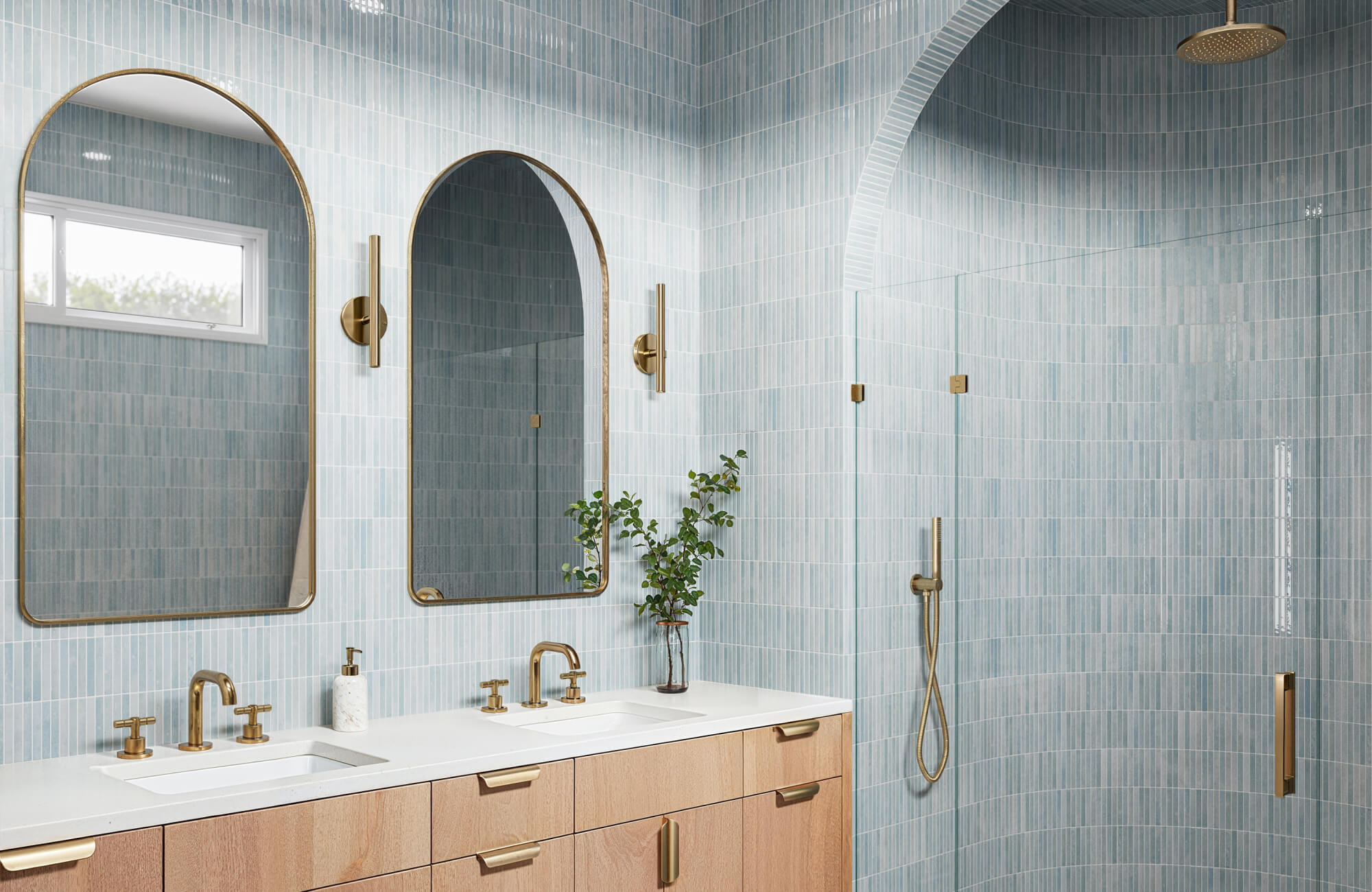 Light blue vertical tiles wrap a contemporary bathroom with arch shower, gold fixtures, and blonde wood vanity.