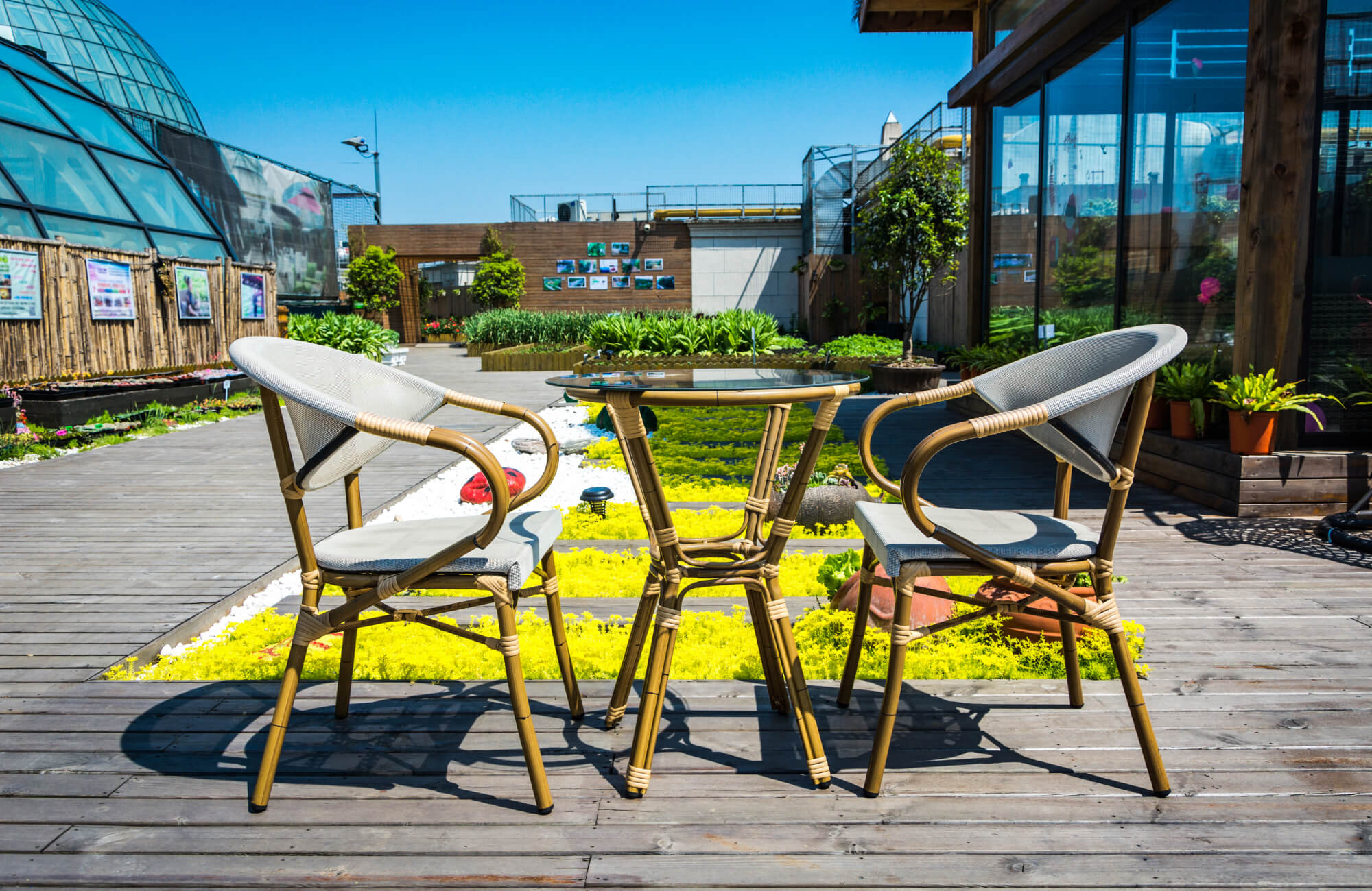 Weather-resistant rattan dining chairs paired with a glass-top table on a sunlit deck, styled within a landscaped rooftop garden with greenery and modern structures.