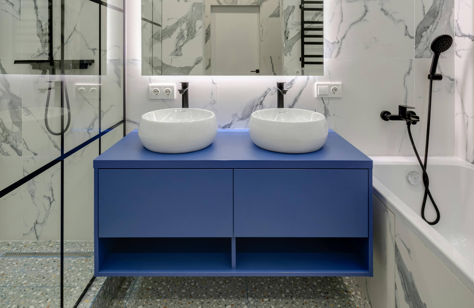 Modern bathroom vanity in cobalt blue with twin round vessel sinks and matte black fixtures, set against marble walls.