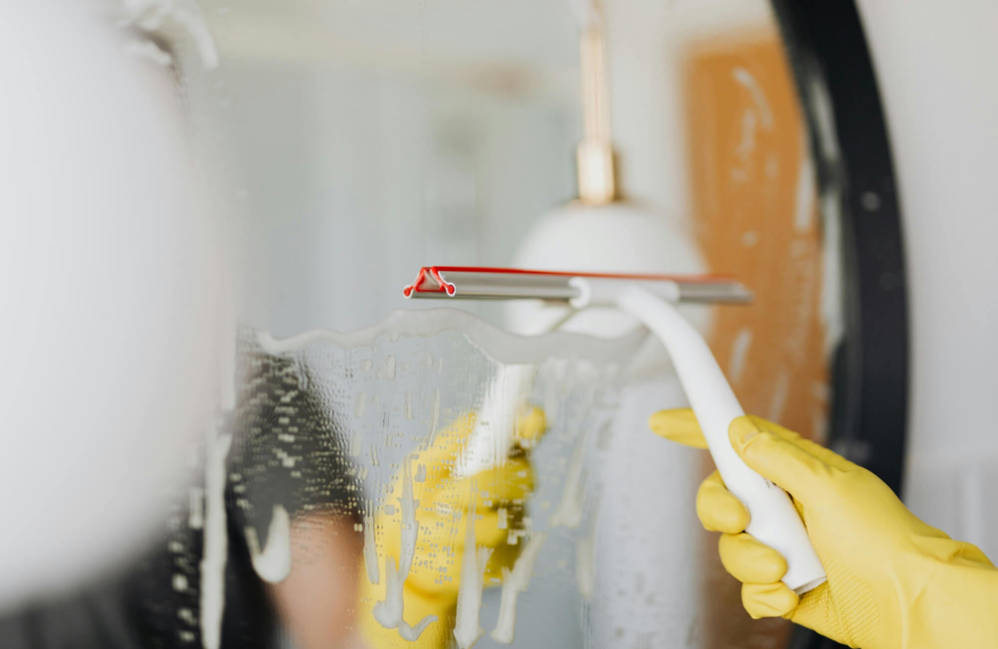 Close-up of a person cleaning a mirror with yellow gloves and a squeegee for a streak-free shine.
