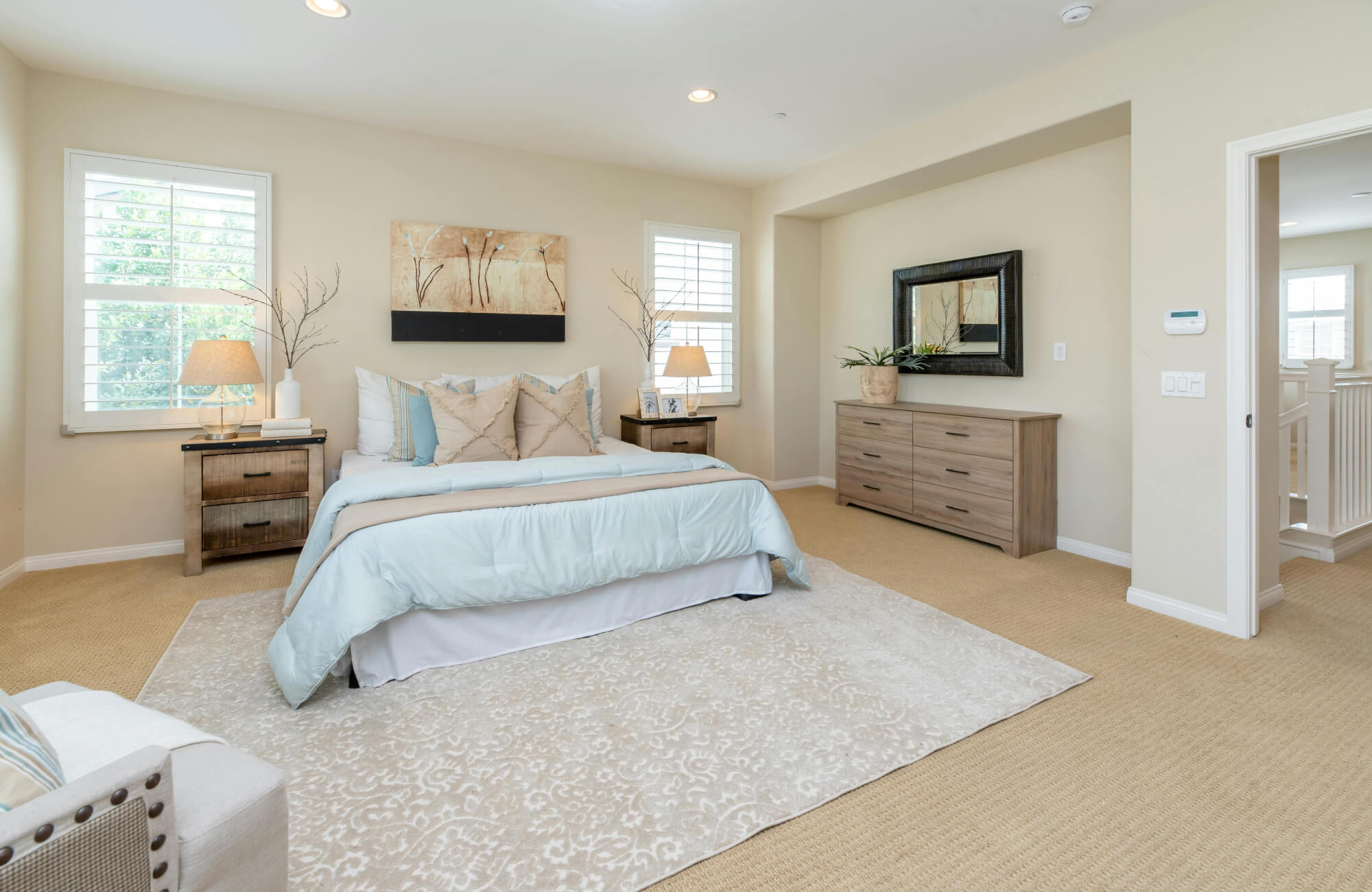 null Spacious beige bedroom with botanical wall art above the bed, pale blue bedding, matching wood nightstands, a large rug, and a framed mirror above a dresser.