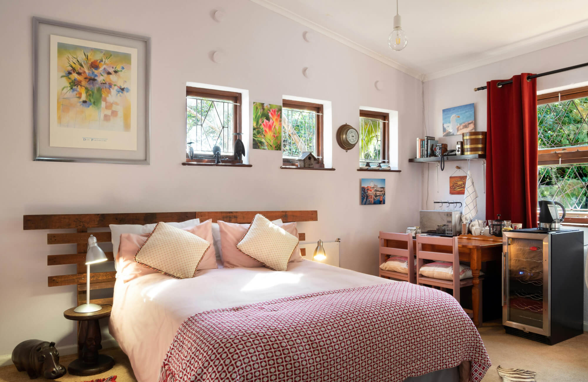 null Cozy bedroom with colorful framed floral art, small wall prints, wood headboard, red patterned blanket, café-style corner setup, and red curtains by the window.