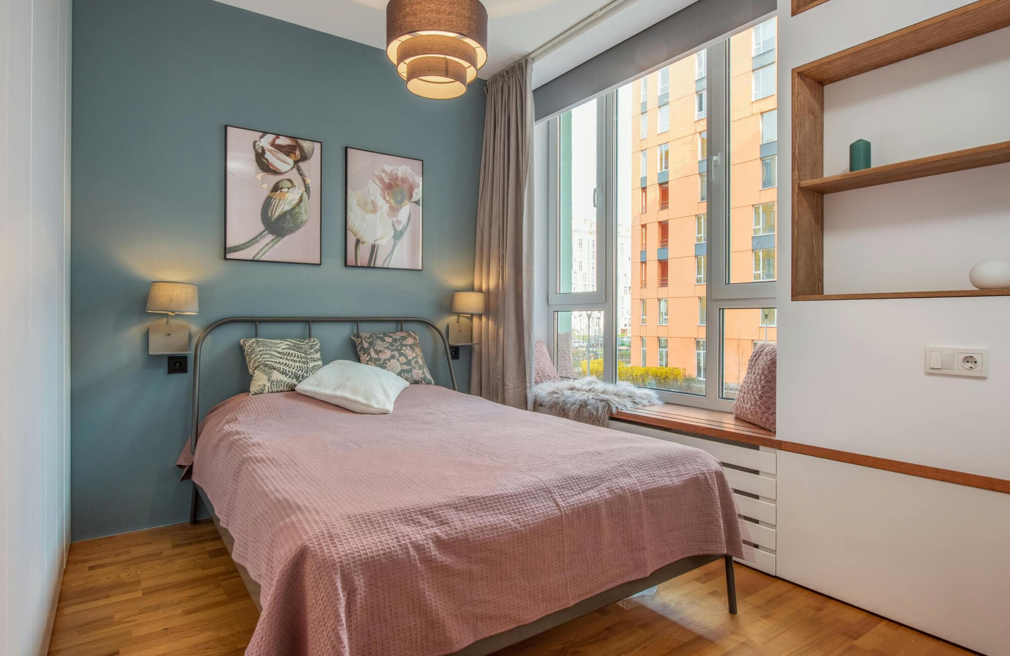 null Compact bedroom with two floral prints above a metal bed, dusty pink bedding, blue-green accent wall, built-in shelving, wood floor, and window bench.