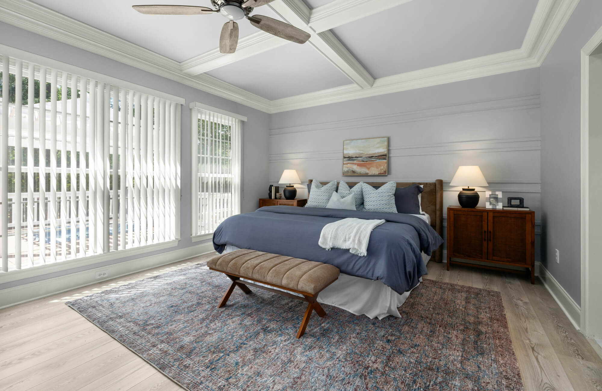 null Traditional bedroom with soft landscape wall art above the bed, blue bedding, matching lamps, tray ceiling, large patterned rug, and tall windows with white blinds.