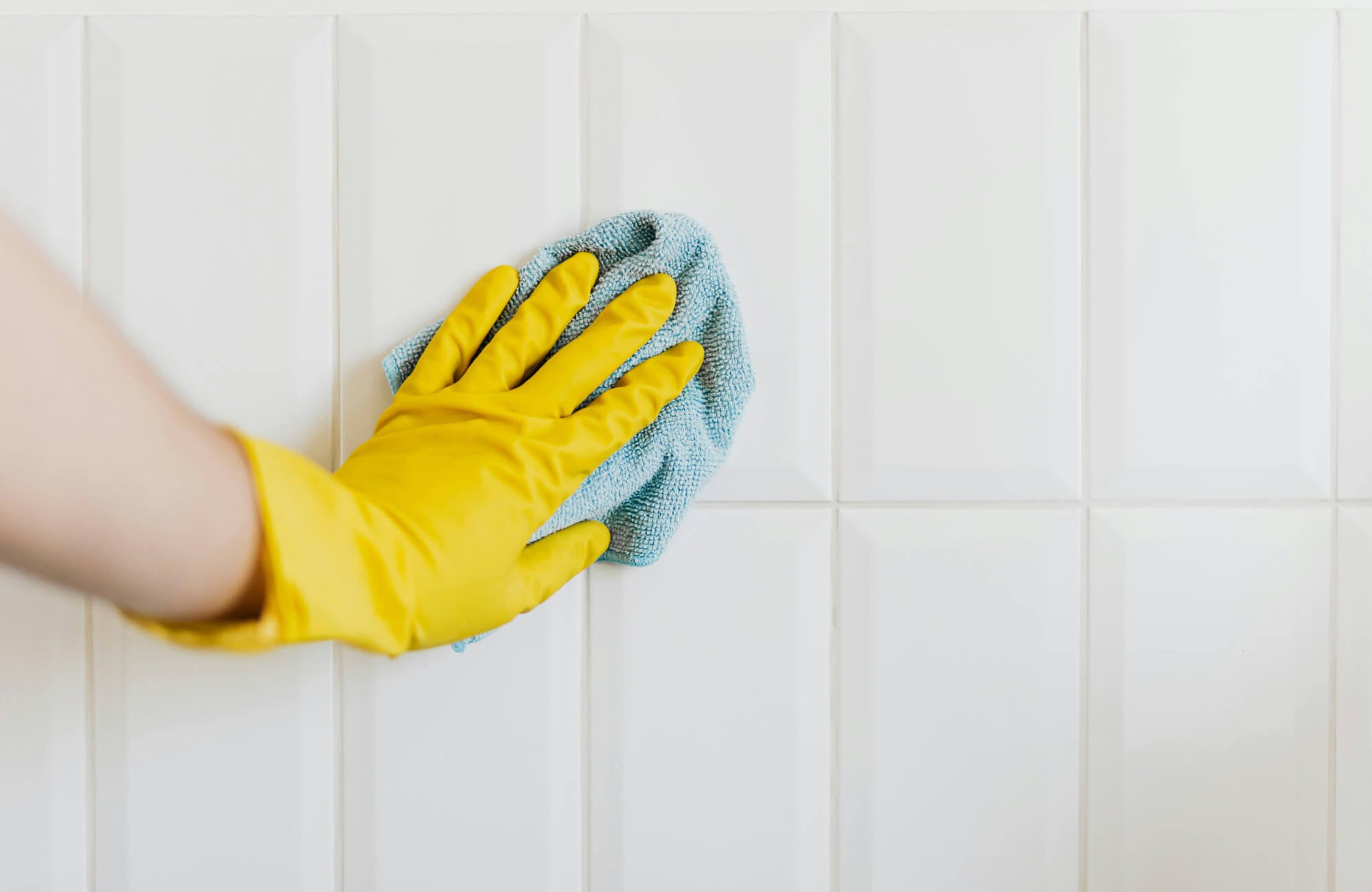 Hand in yellow glove wiping glossy white ceramic wall tiles with blue microfiber cloth for a sparkling clean bathroom look and easy tile maintenance