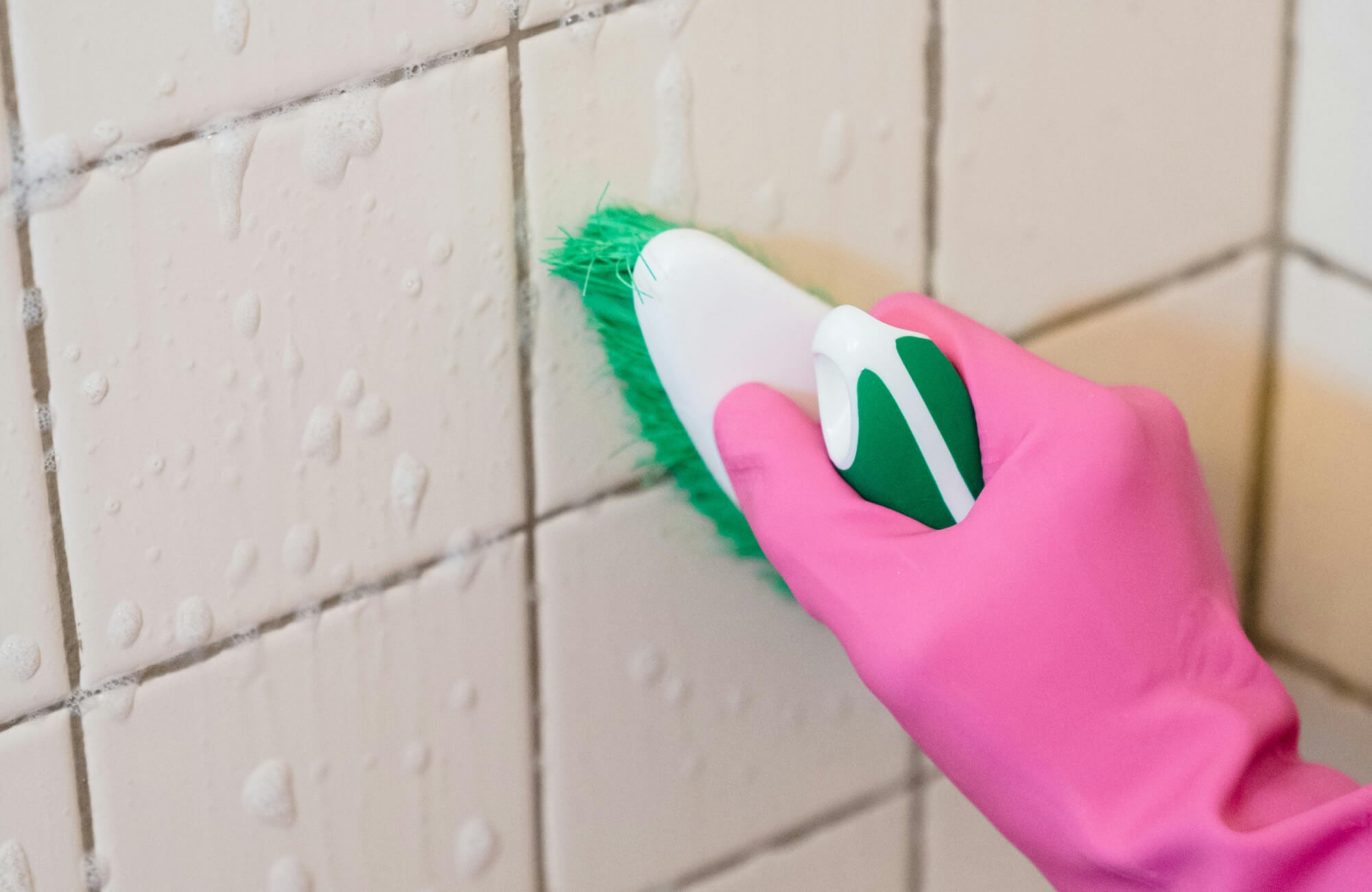 Close-up of pink-gloved hand scrubbing soapy white ceramic tiles with green brush for deep grout cleaning and tile refresh in bathroom