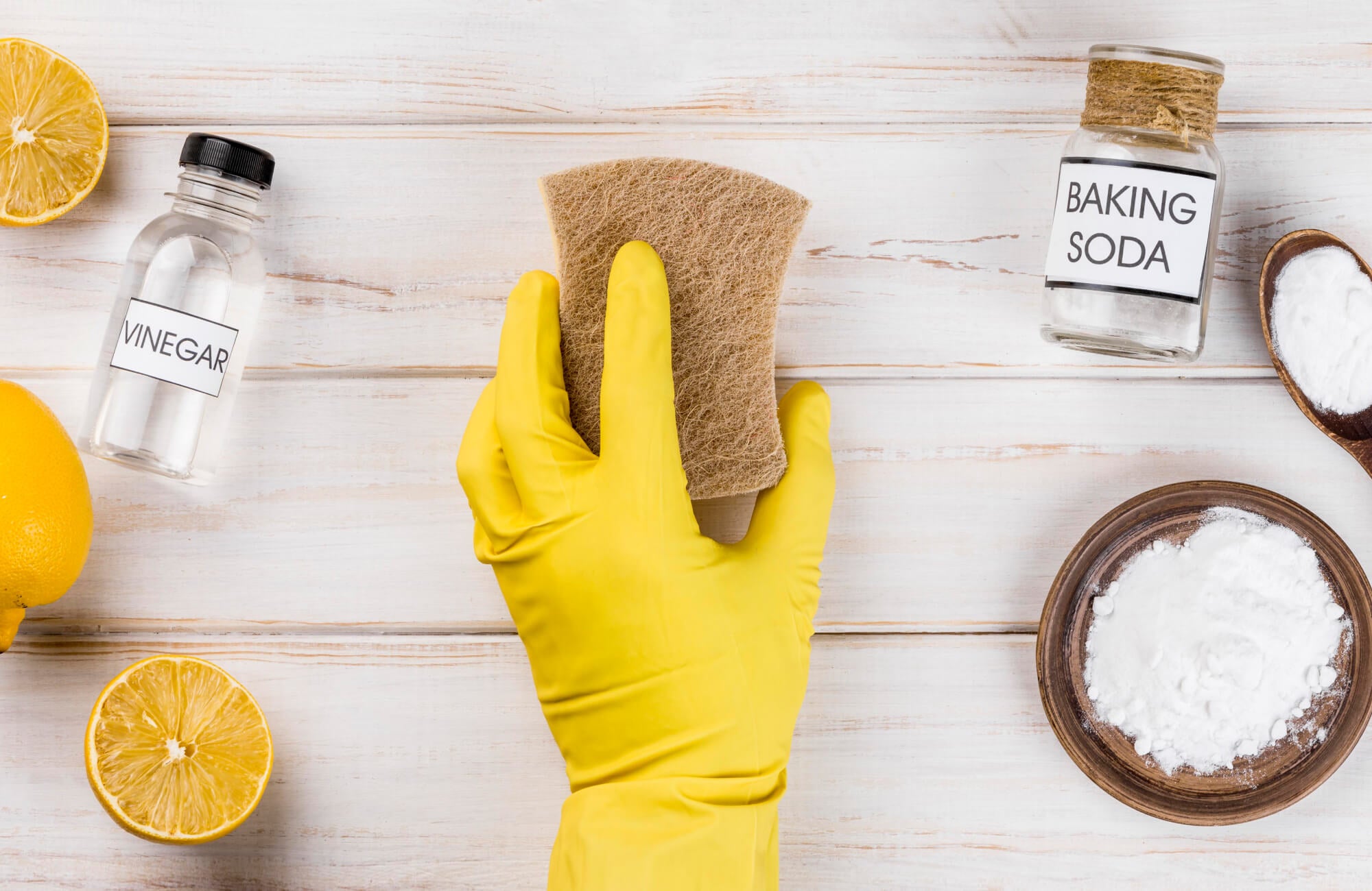 Natural tile cleaning setup with vinegar, baking soda, lemon halves, and a scrubber in a yellow glove on rustic white wood background