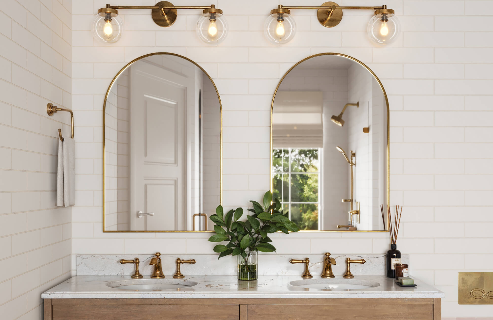 Elegant bathroom with glossy white subway tiles, arched gold-framed mirrors, and marble countertops, complemented by warm brass fixtures and natural light.