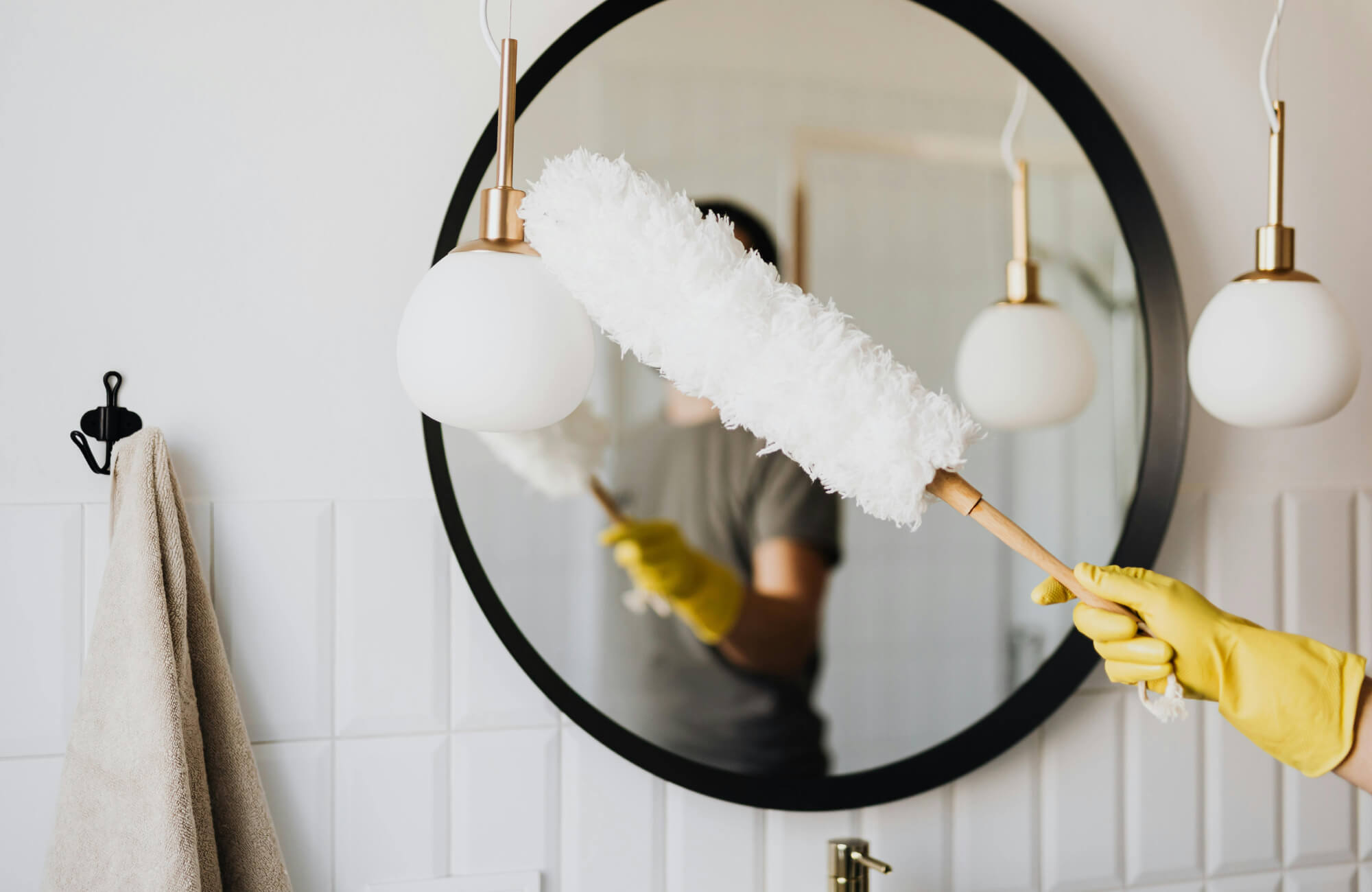Bathroom mirror being dusted with a soft white duster, featuring gold-accented globe pendant lights and a beige towel hanging nearby.