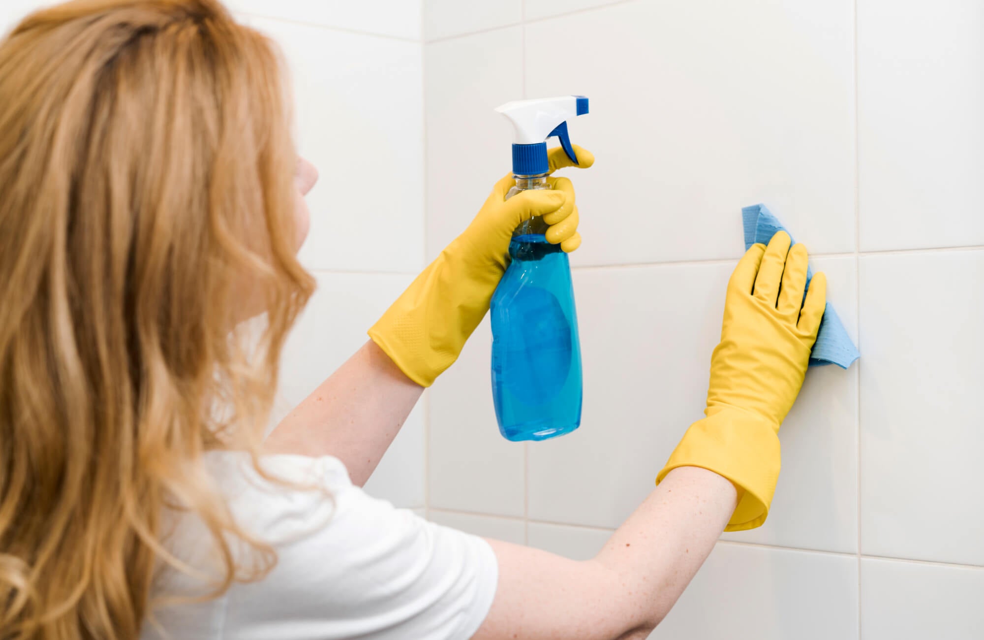 Close-up of tile cleaning in a bathroom, showing gloved hands wiping the surface with a cloth and cleaning solution.