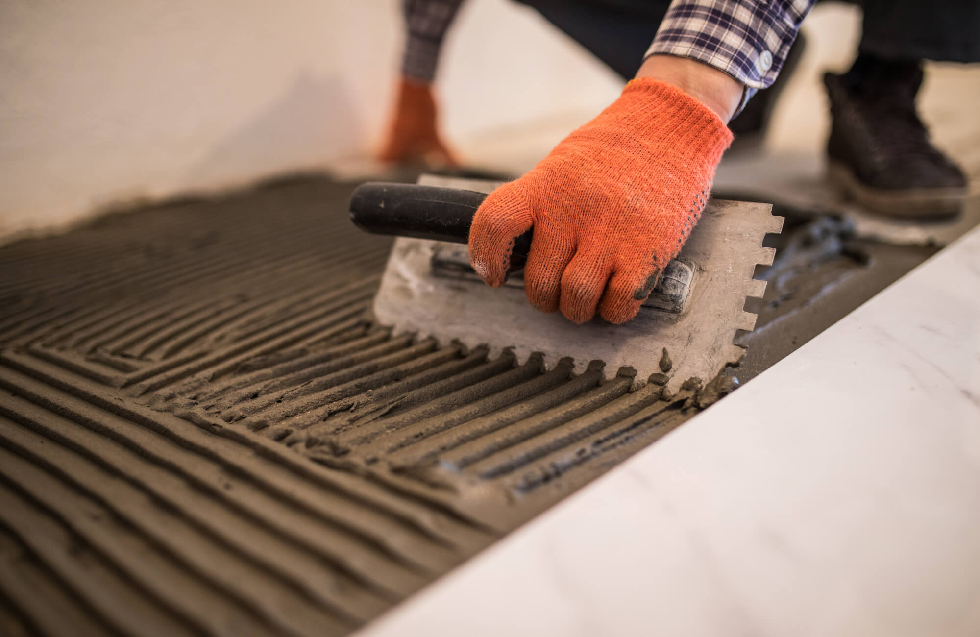 Close-up of hands spreading thinset mortar on subfloor with a notched trowel during tile installation.