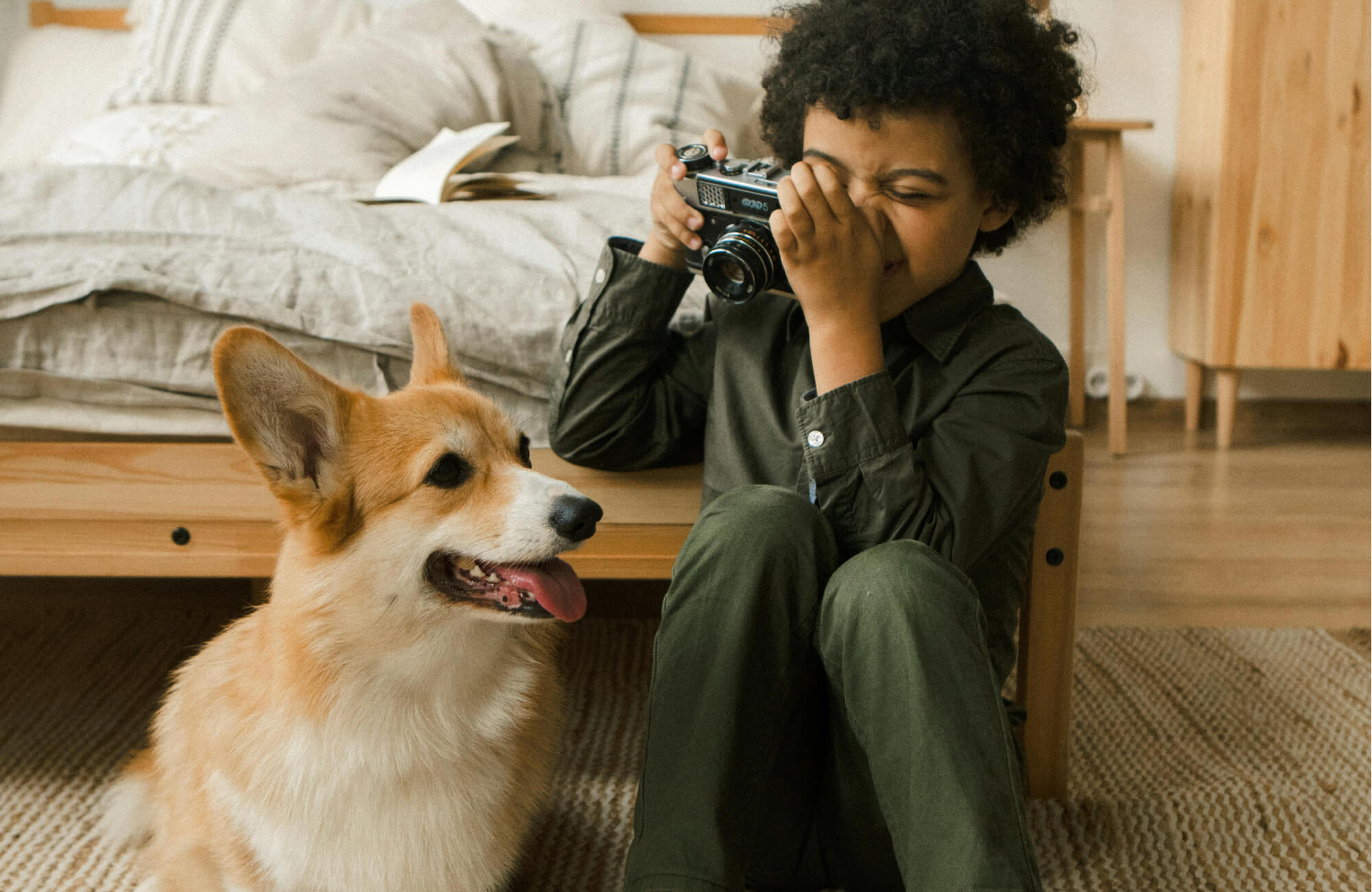 null A smiling child takes a photo with a vintage camera beside a happy corgi on a textured woven rug in a cozy bedroom.