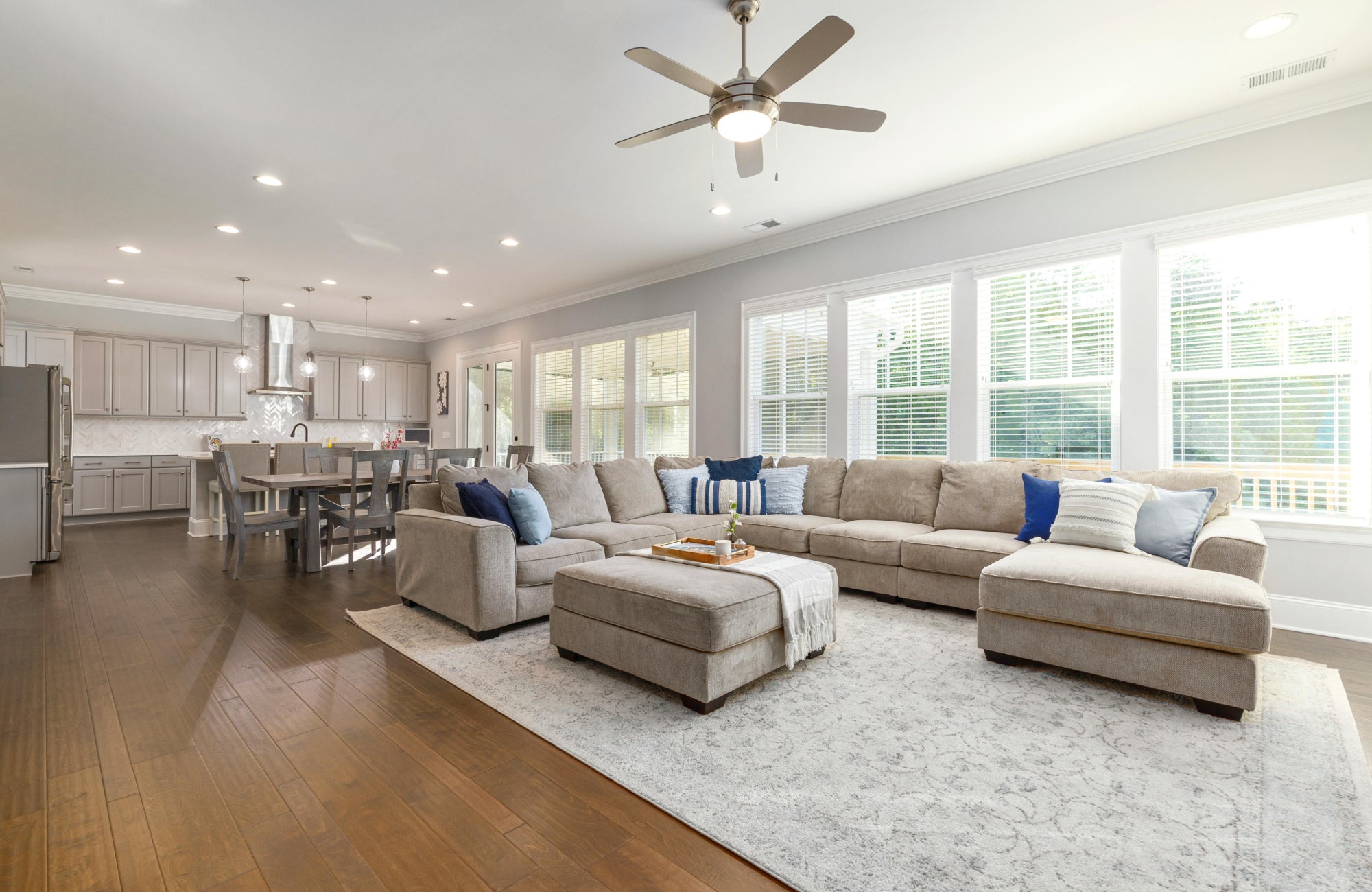 Spacious open-plan living area with soft gray sectional, modern ceiling fan, and kitchen island illuminated by pendant lights, all flooded with daylight from large windows.