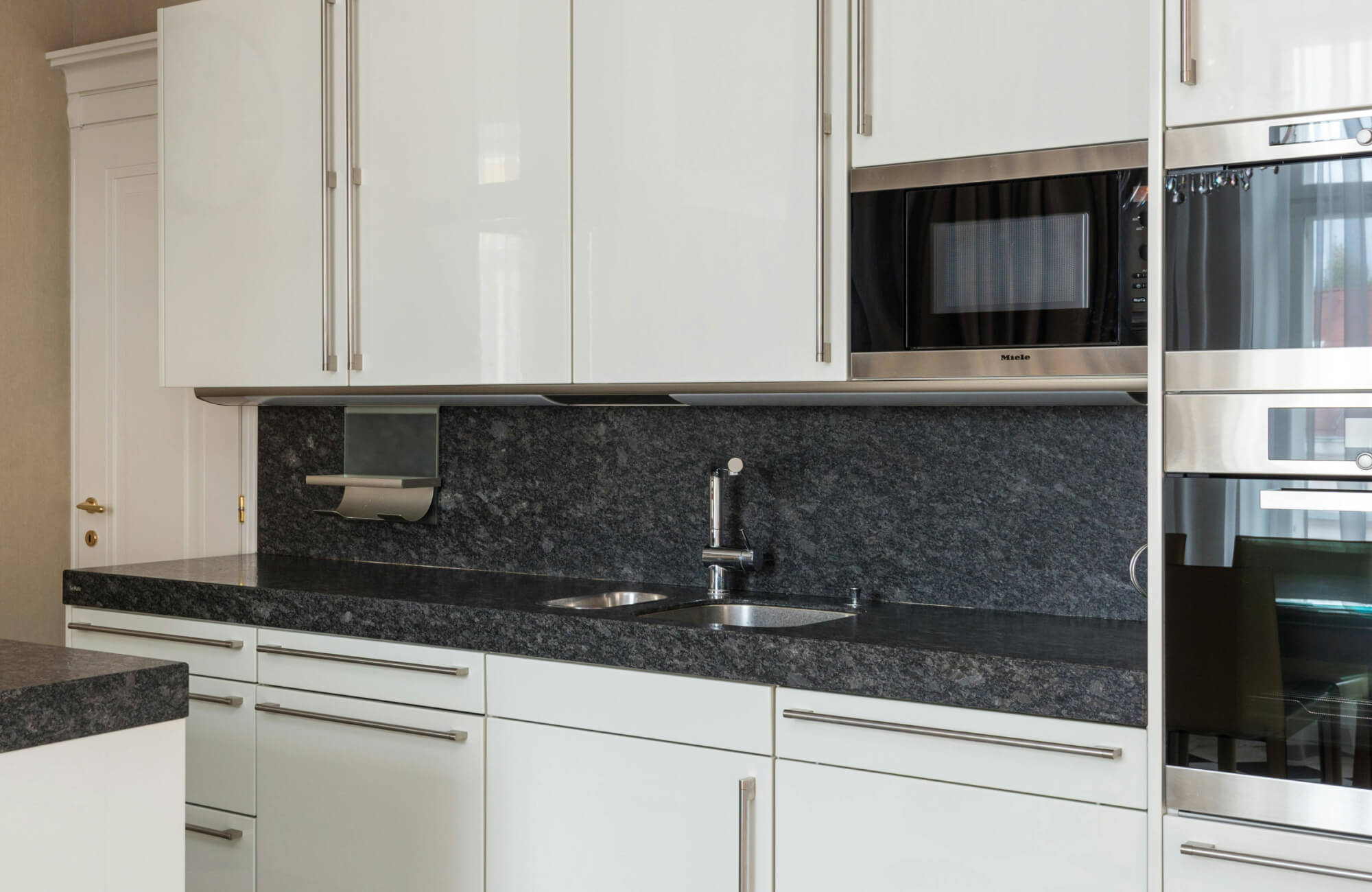 null Sleek white cabinetry with a matching dark stone backsplash and counter, featuring a modern dual sink and integrated microwave and oven.