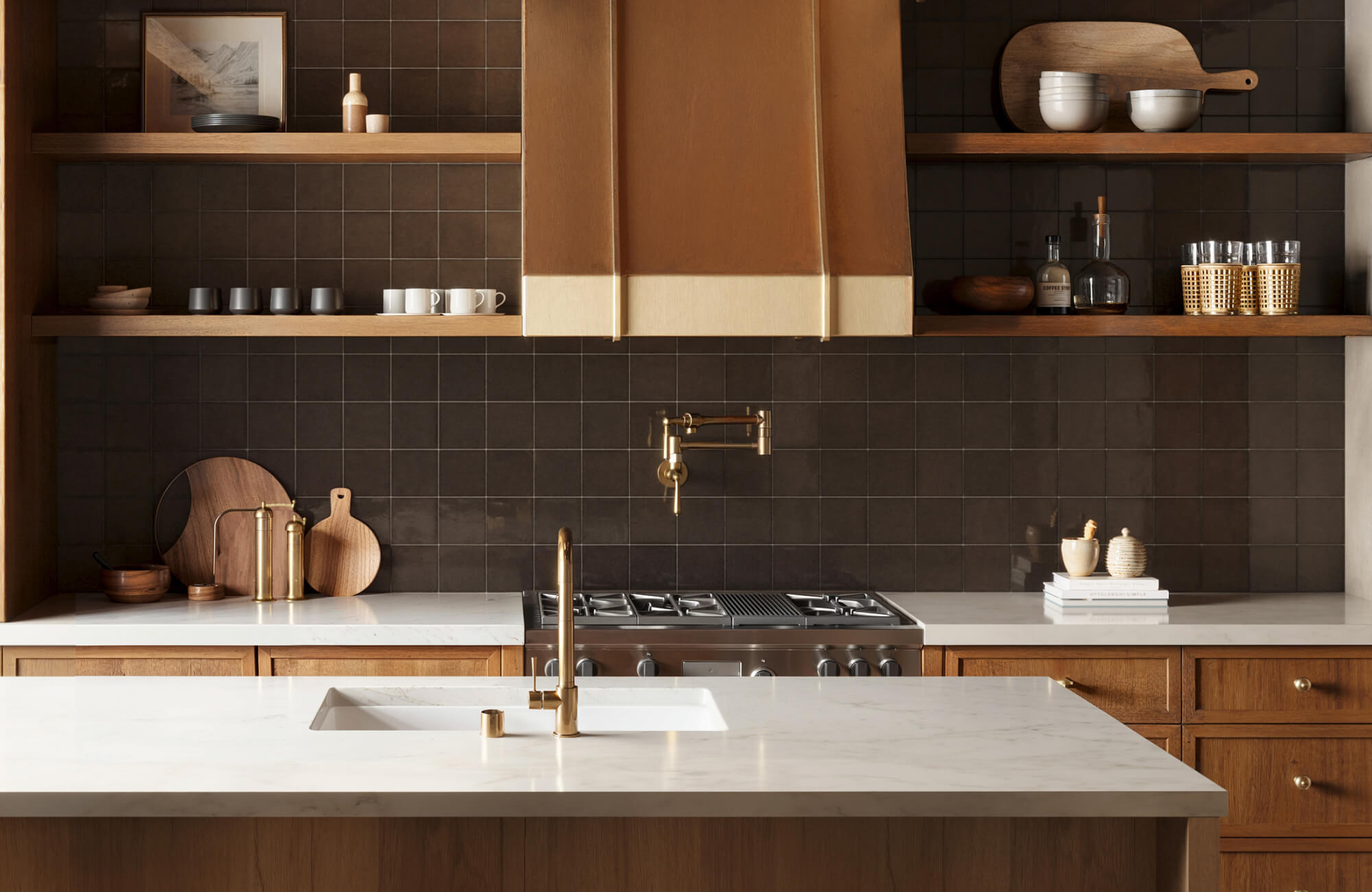 null Warm-toned kitchen design with dark backsplash tiles, marble countertops, and a gold range hood paired with open wooden shelves and neutral decor.