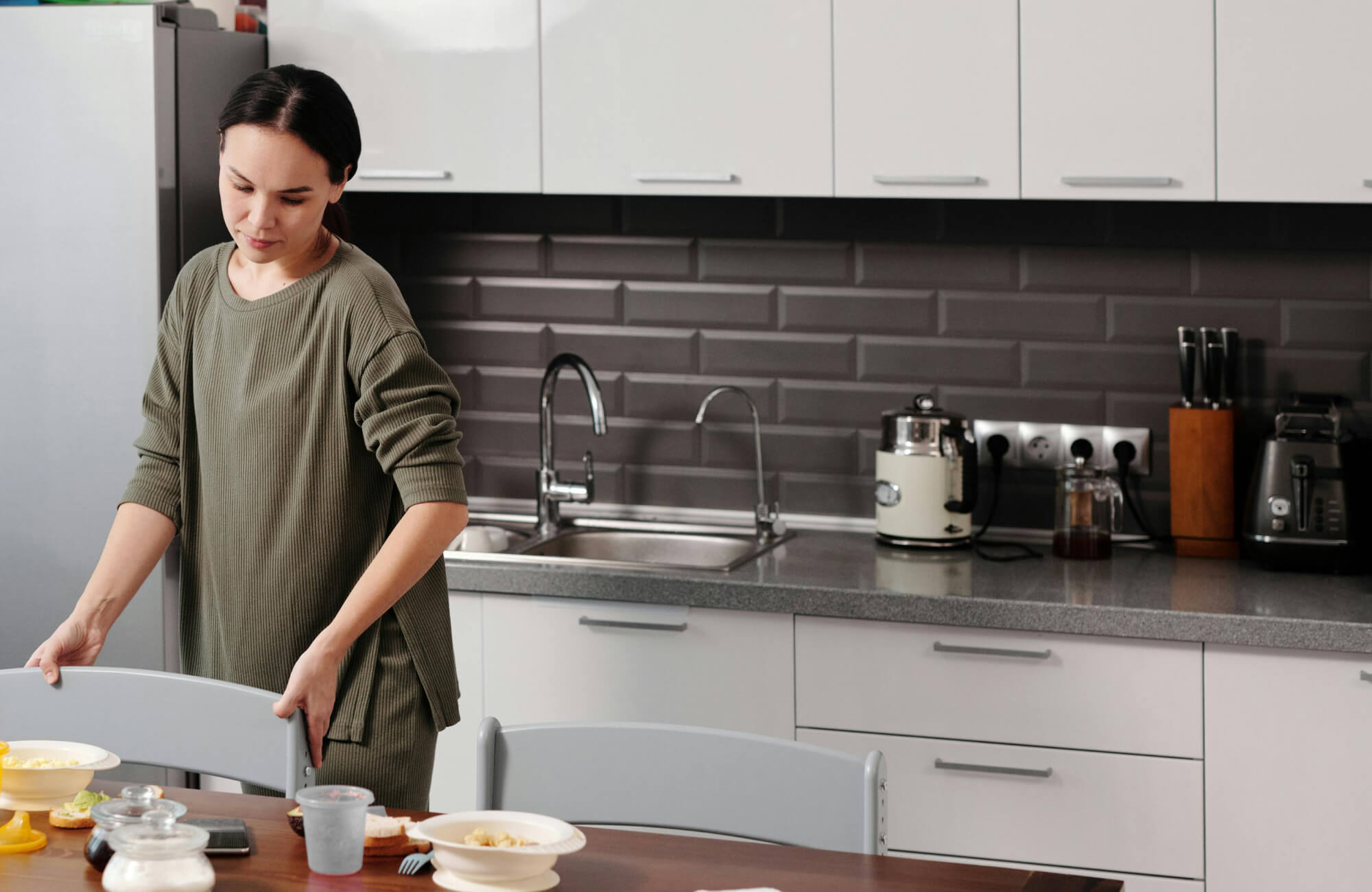 null Modern kitchen featuring raised rectangular gray tiles, double sink, silver appliances, and a casual dining setup with a person tidying up.