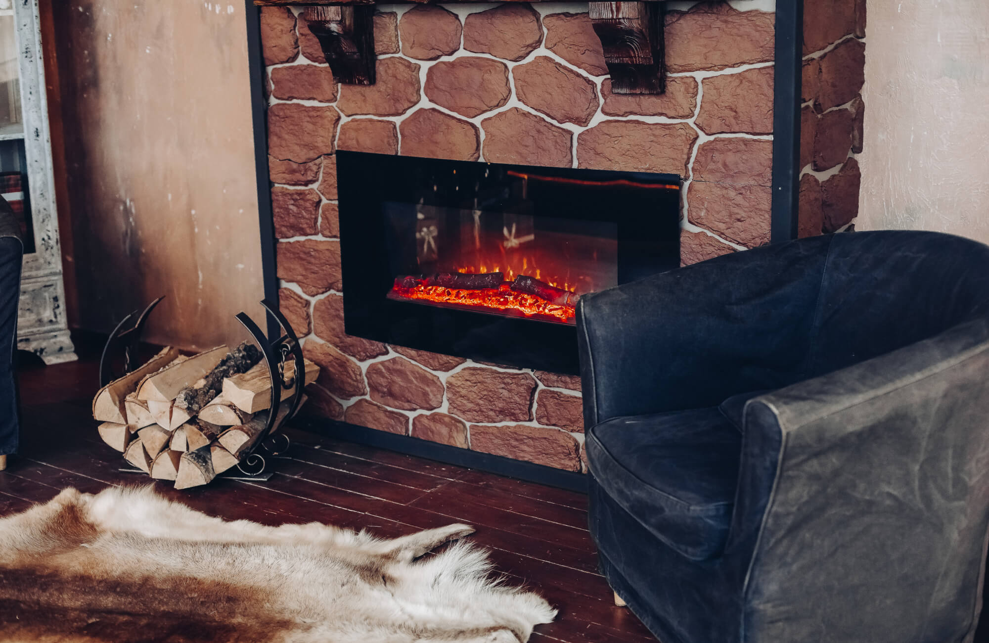 Cozy living space featuring a stone-clad fireplace with an electric flame insert, a dark velvet armchair, and firewood accents that highlight the rustic charm.