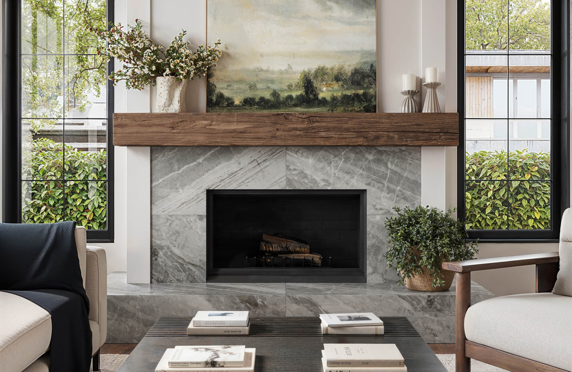 A sleek fireplace surrounded by gray marble tiles and topped with a warm wood mantel decorated with flowers and candles, set between large black-framed windows.