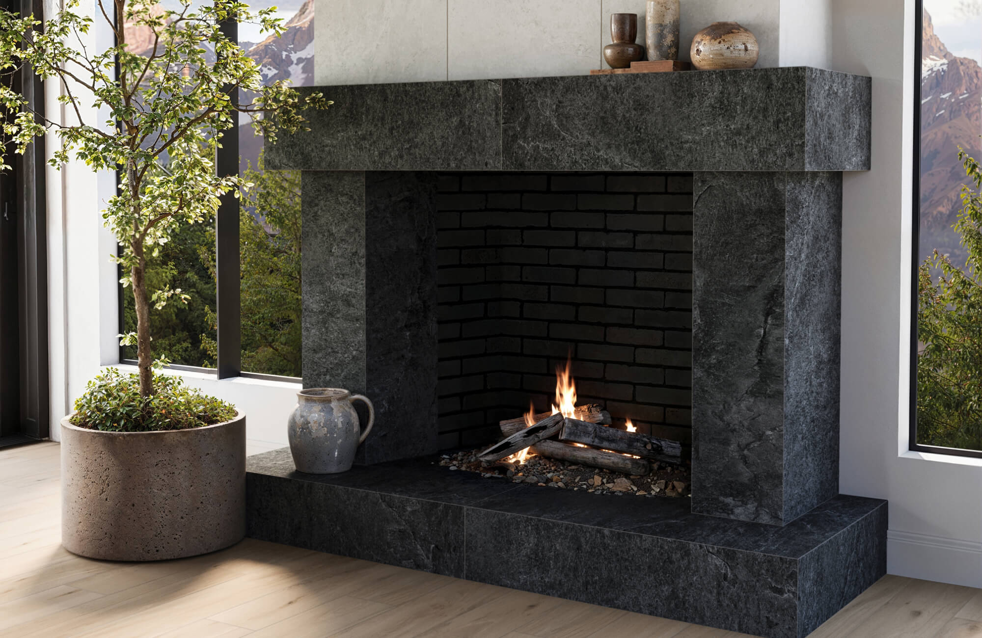 A modern fireplace framed in dark stone with a burning fire inside, styled with rustic pottery and set beside large windows overlooking a mountain landscape.