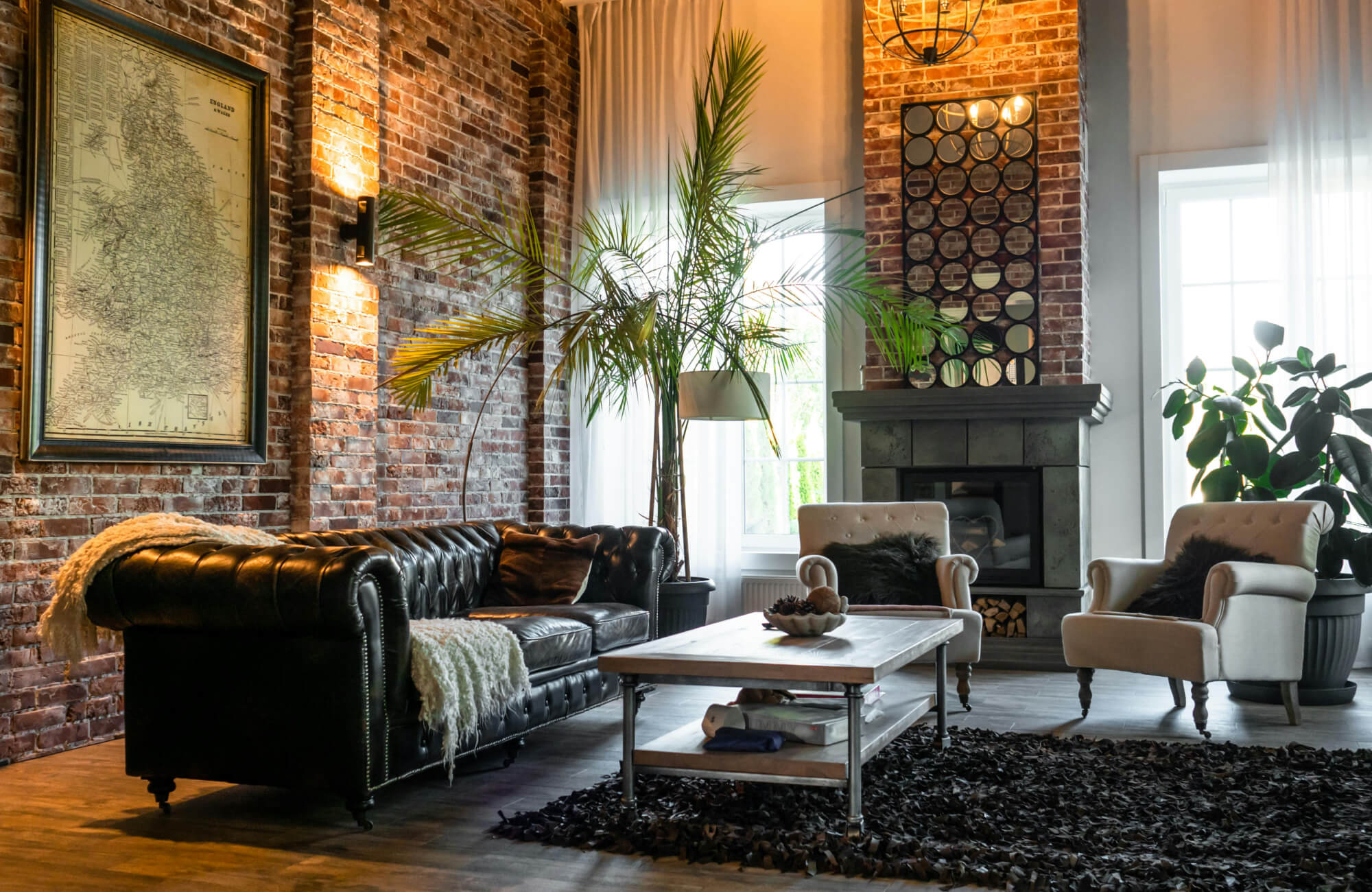 A cozy industrial style living room with exposed brick walls, a dark leather sofa, soft armchairs, and a stone fireplace framed by tall windows and indoor greenery.