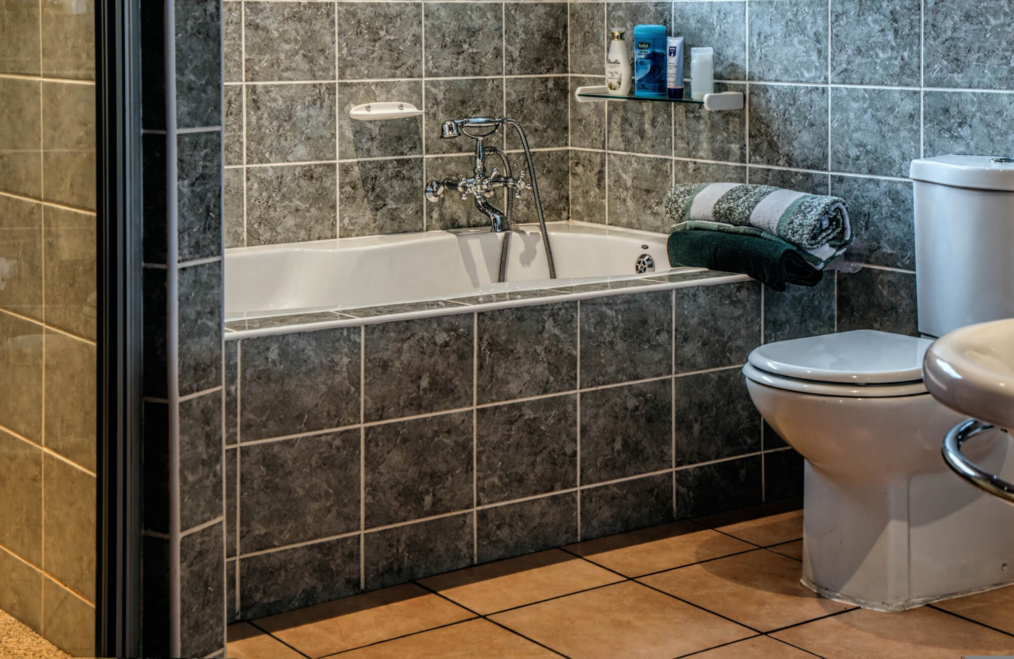 Modern bathroom featuring dark gray tiled walls, a white bathtub, and a stained tiled floor, with towels on display.