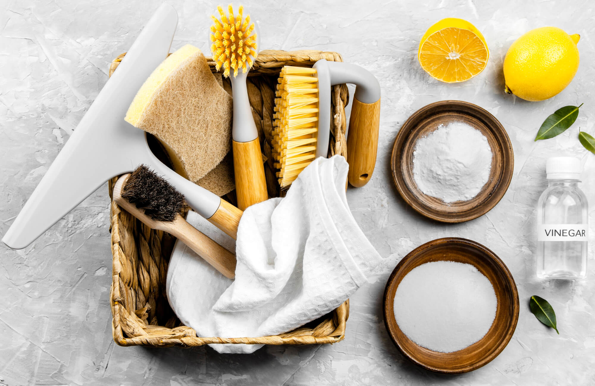 Natural cleaning kit with brushes, sponges, lemon, vinegar, and baking soda arranged in a woven basket on a gray surface.