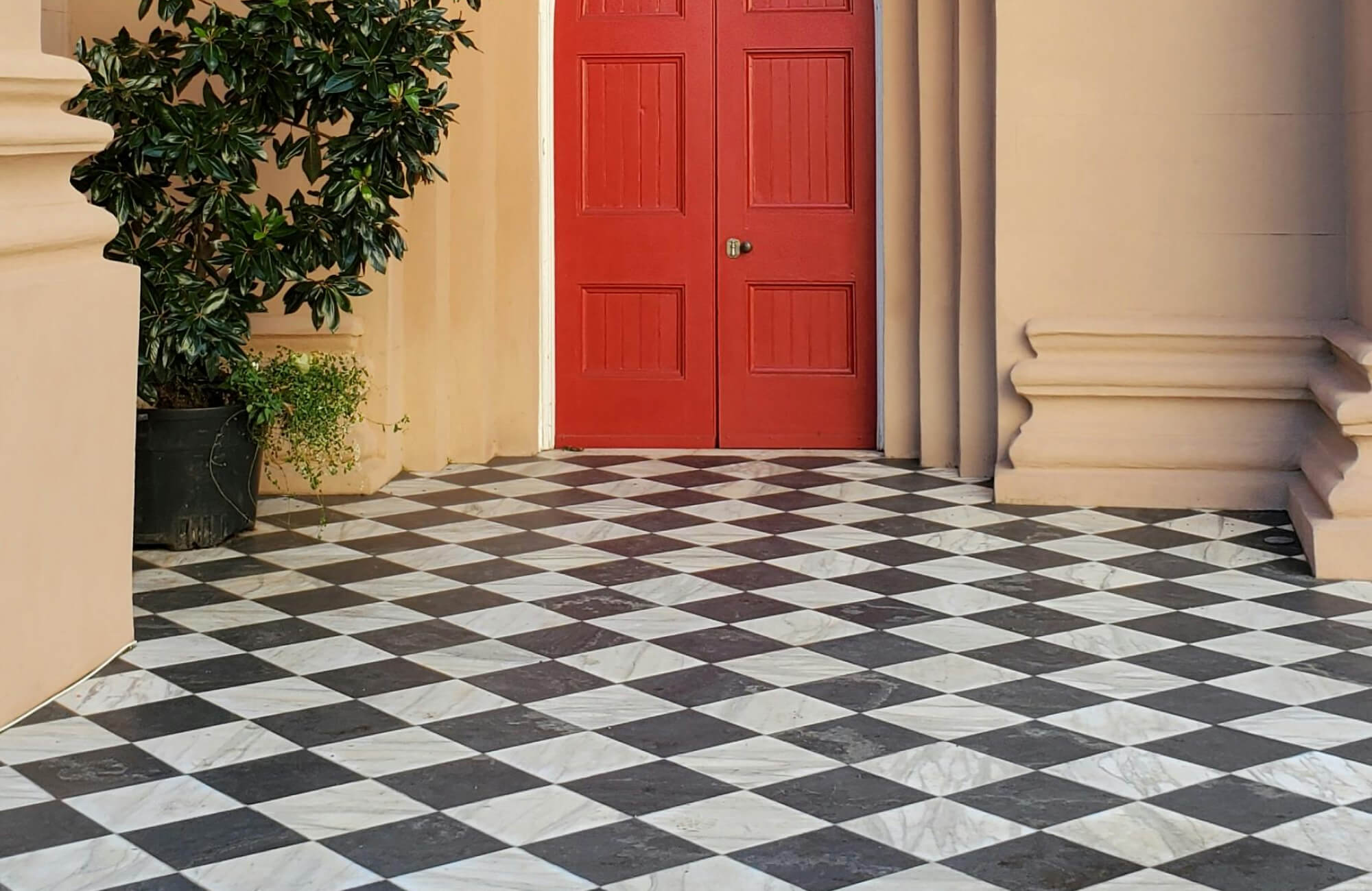 null Black and white marble checkerboard tiles enhance the entrance, contrasting elegantly with vibrant red double doors and neutral building tones.