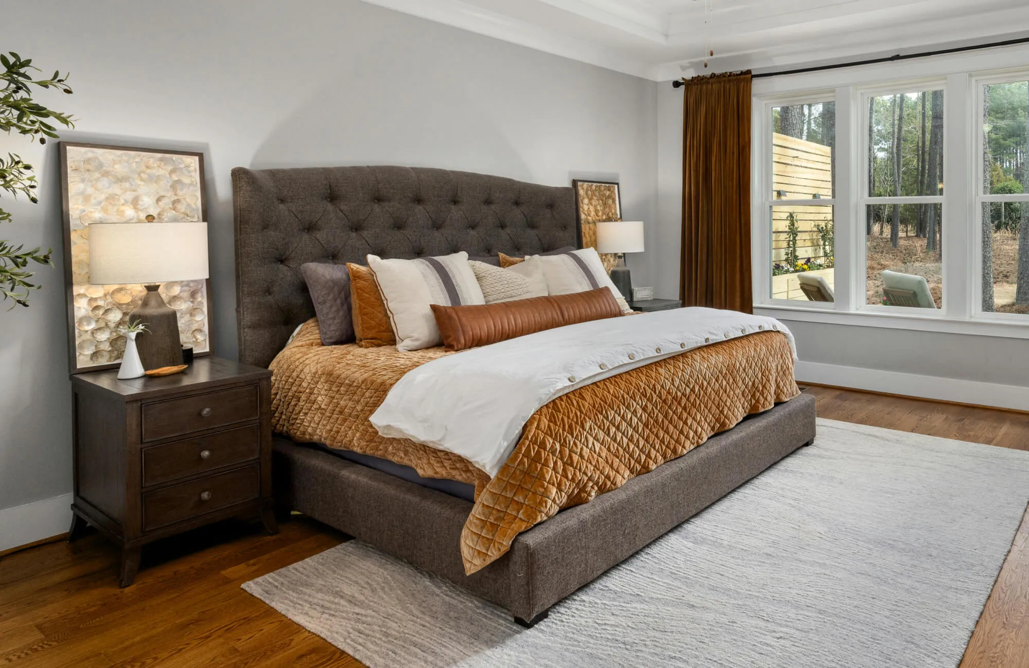 Neutral bedroom with a tufted gray headboard, rust quilted bedding, layered pillows, and a long brown bolster pillow centered across the bed.
