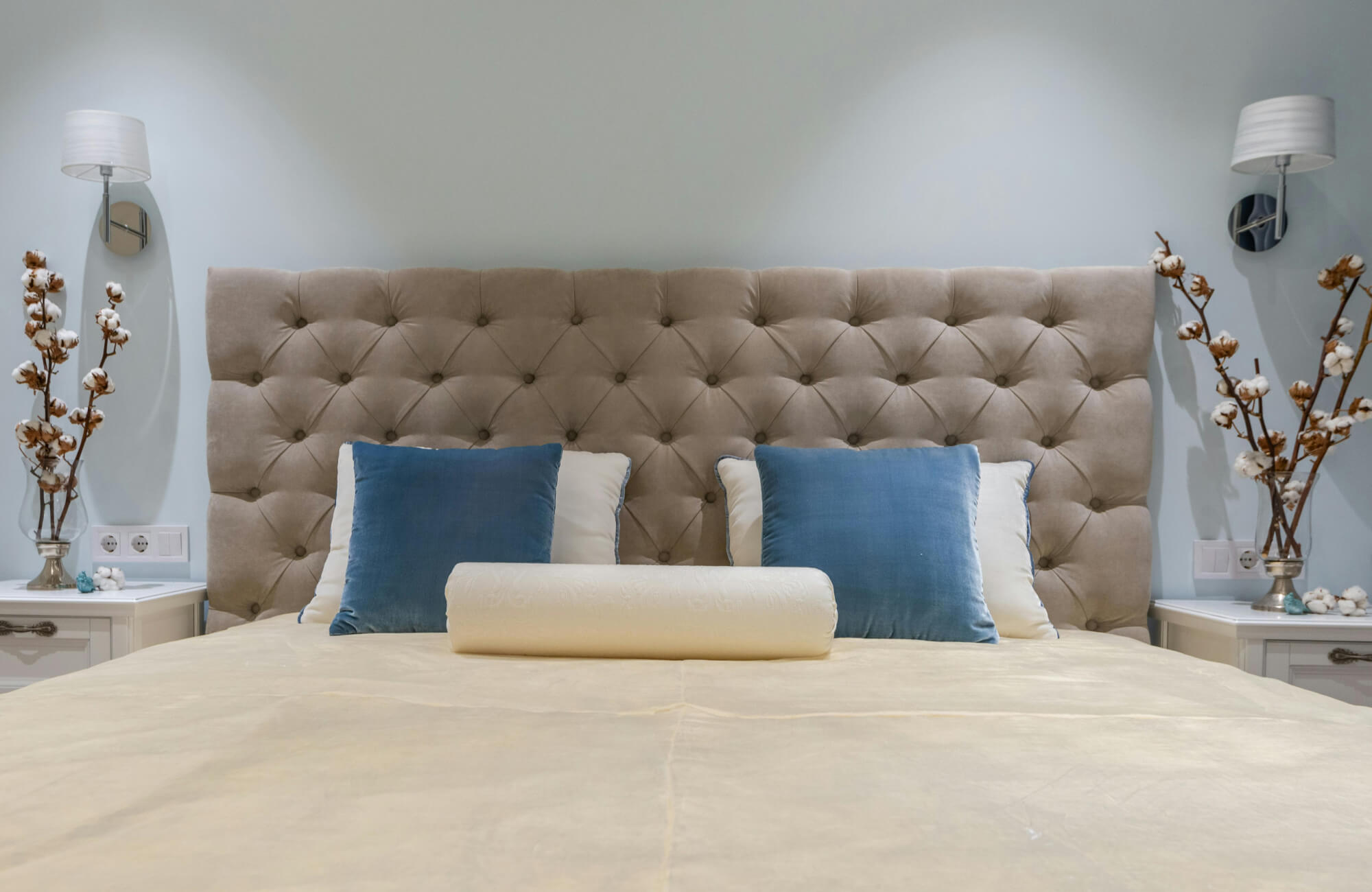 null Beige bedroom with a tufted upholstered headboard, blue accent pillows, and a cream cylindrical bolster pillow placed at the front of the bed.