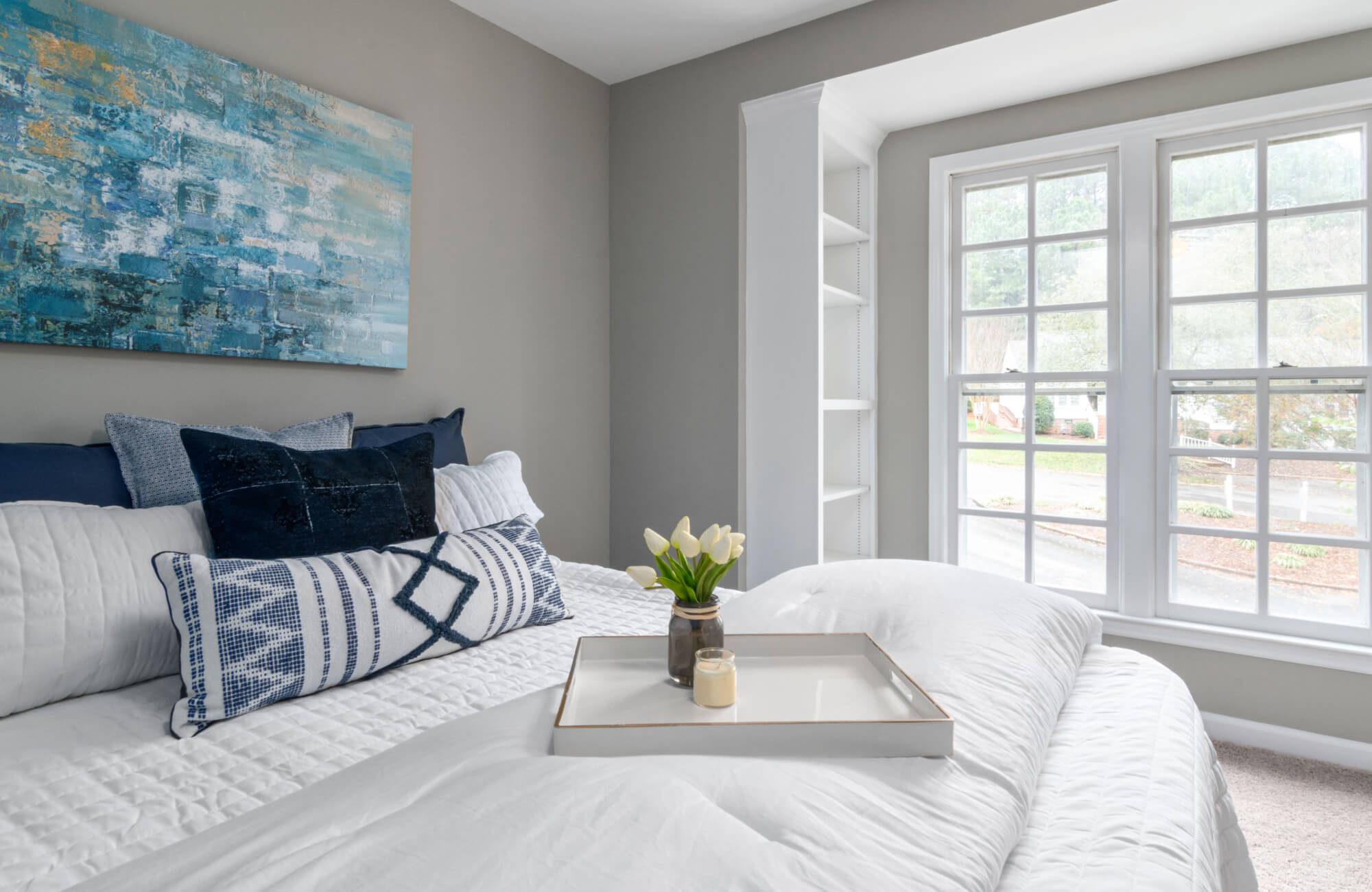 null Bright bedroom with white bedding, layered blue accent pillows, a patterned bolster pillow, large windows, and abstract blue wall art above the bed.