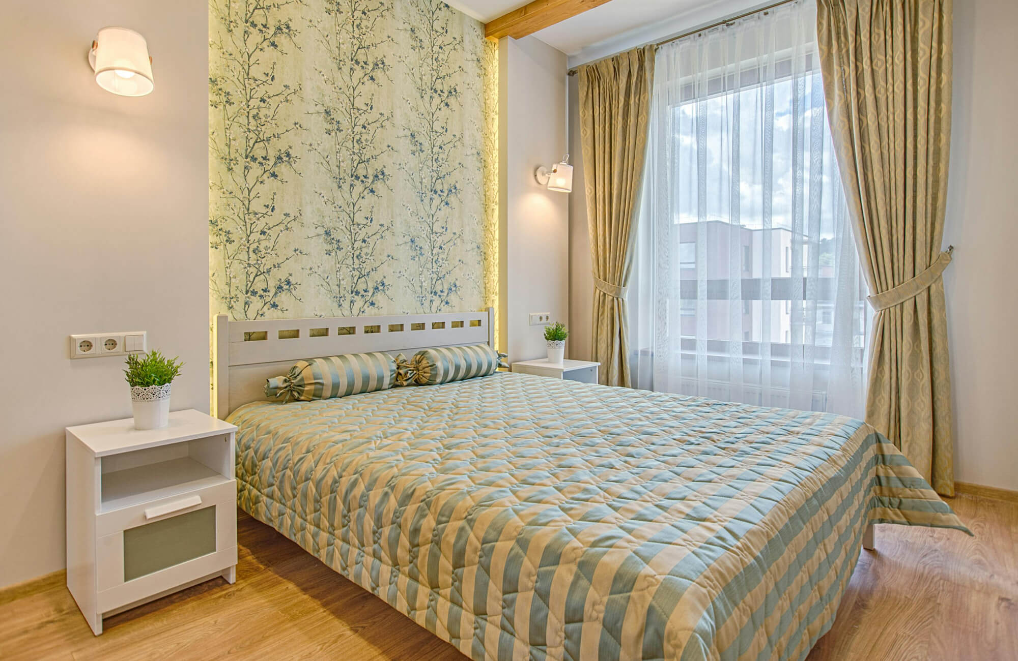 null Soft-toned bedroom with striped blue and beige bedding, matching cylindrical bolster pillows, floral wallpaper, and full-length curtains.
