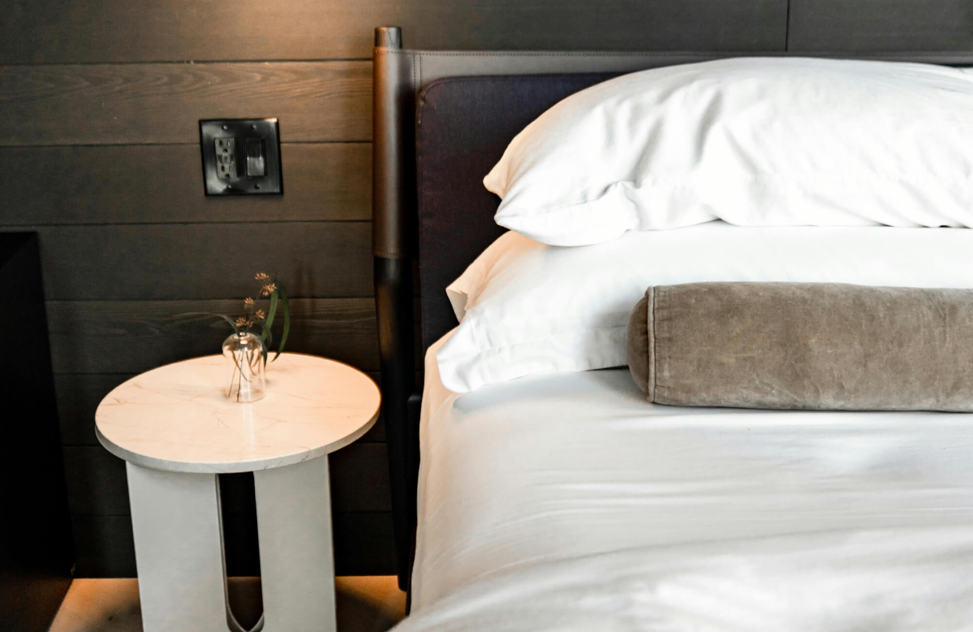 null Close-up of a modern bed with white bedding, stacked sleeping pillows, a gray cylindrical bolster pillow, and a dark wood-paneled wall.