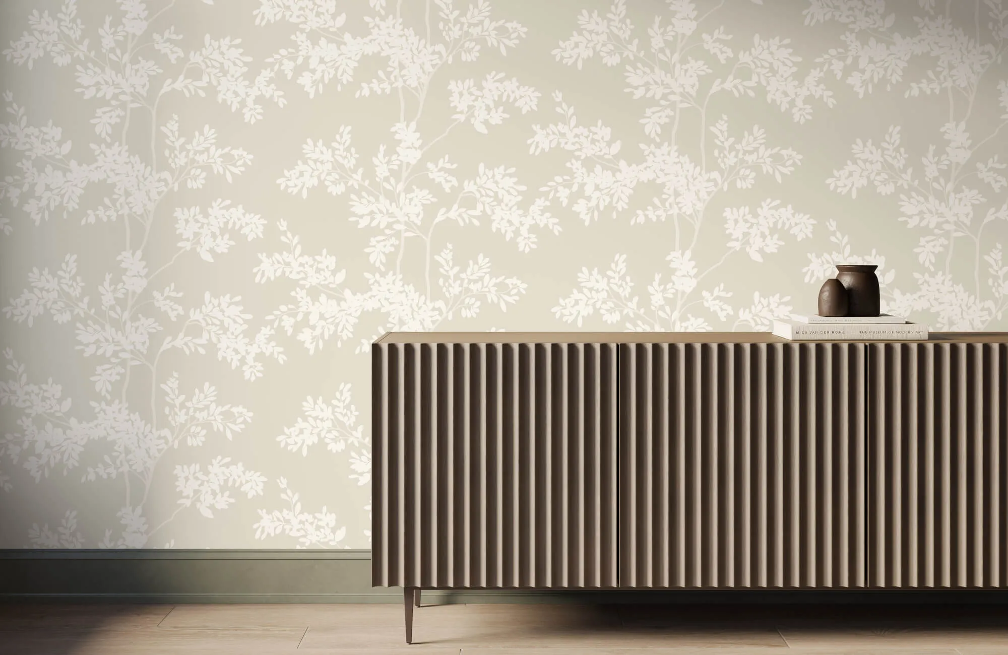 A beige fluted sideboard topped with a dark brown clay vase and stacked books sits against a soft beige wallpaper with a white botanical branch pattern.