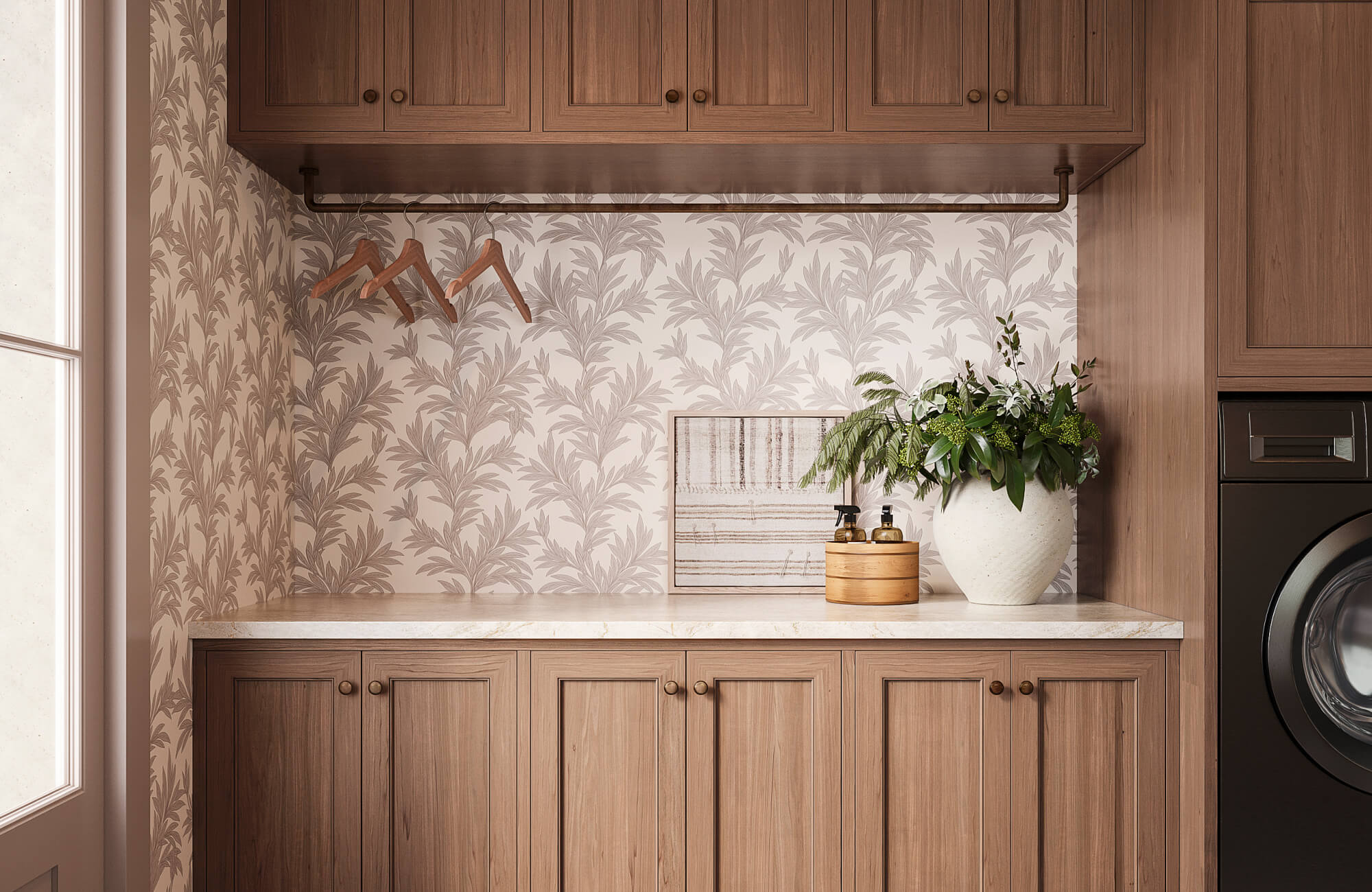 null Warm brown wood upper and lower cabinets frame a cream and gray leaf print wallpaper in a styled laundry room with a white stone countertop and green floral arrangement.