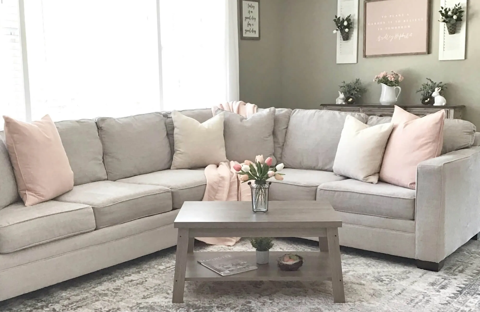 Cozy cottage living room with beige sectional, pink throw pillows, gray coffee table, floral accents, and soft neutral decor.