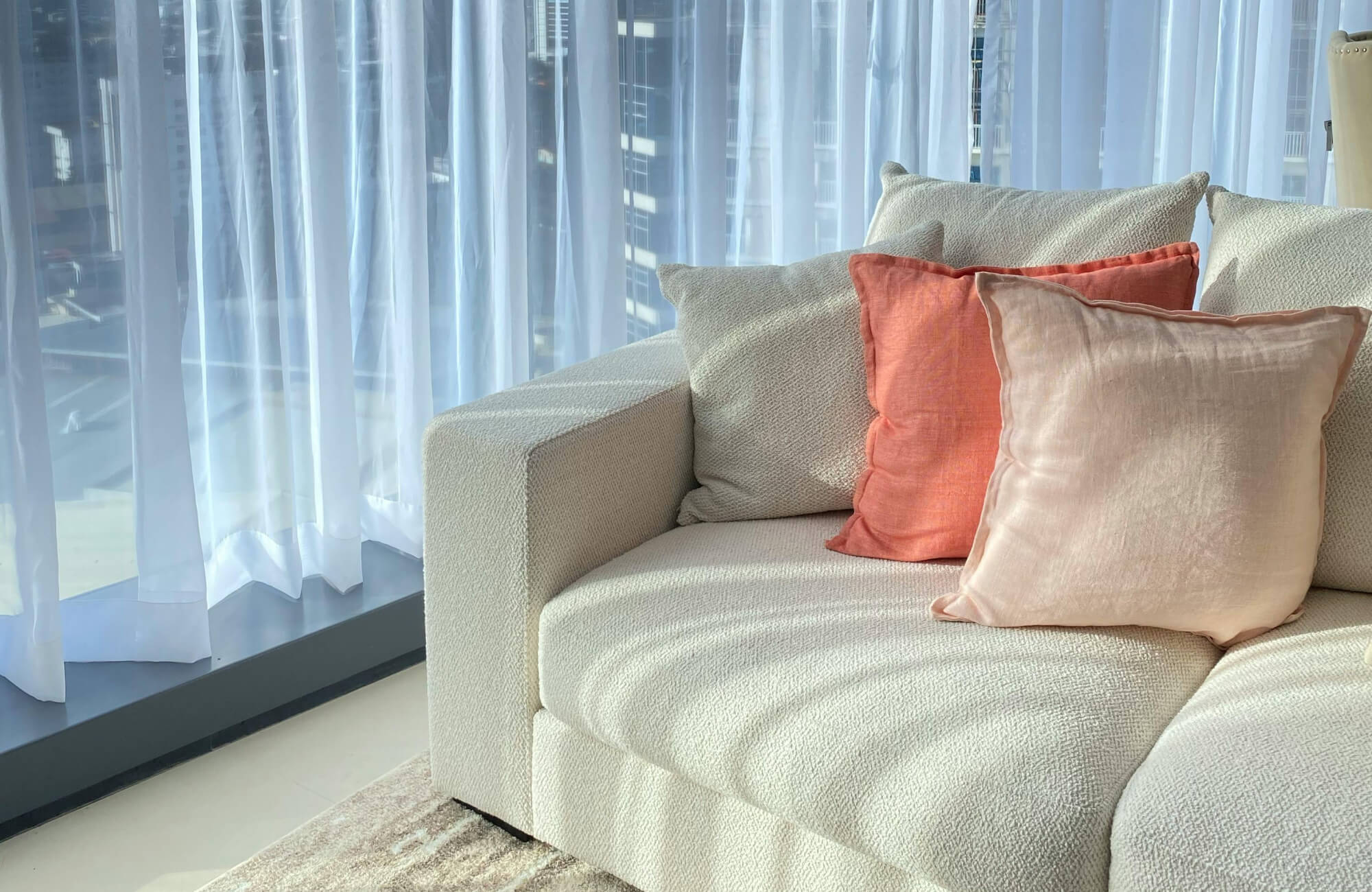 null Modern coastal living room with cream textured sofa, coral pink throw pillows, sheer curtains, and soft city light.