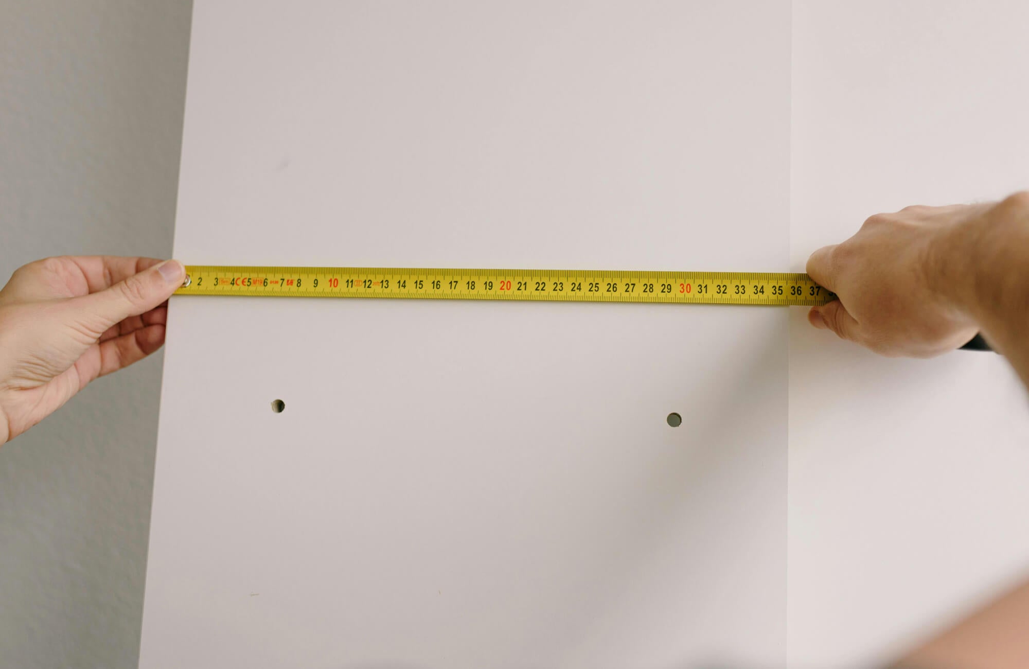 null Person measuring wall width with a tape measure before hanging artwork on a light-colored interior wall with visible mounting holes