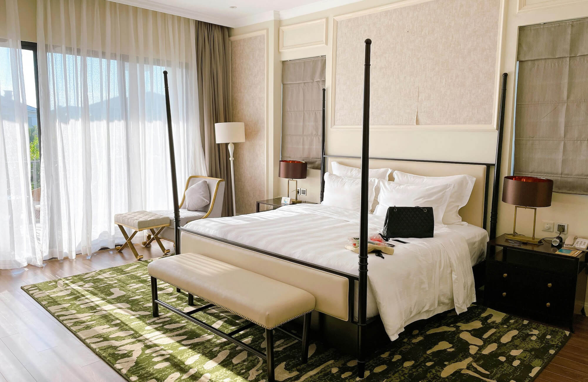 null A bright bedroom with a four-poster bed, layered white bedding, soft neutral tones, and a patterned green rug adding contrast near the large window draped in sheer curtains.