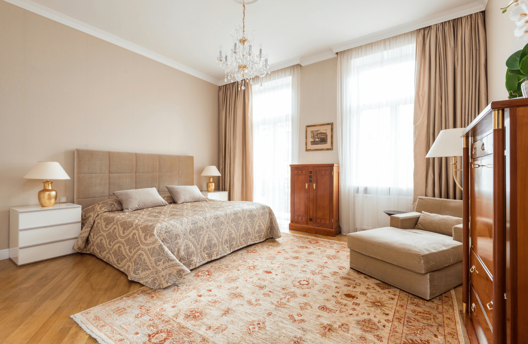 null A spacious beige bedroom featuring a patterned area rug under a low platform bed, with warm wood furniture and soft natural light from tall windows.