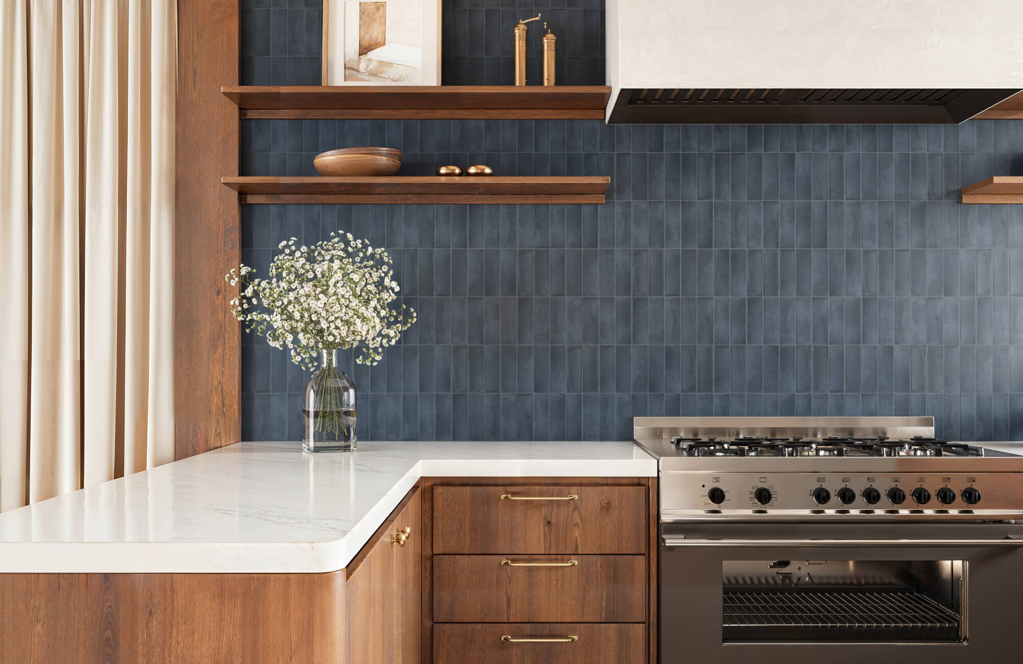 Warm kitchen with vertical blue backsplash tiles, wood cabinetry, floating shelves, and a stainless steel range.
