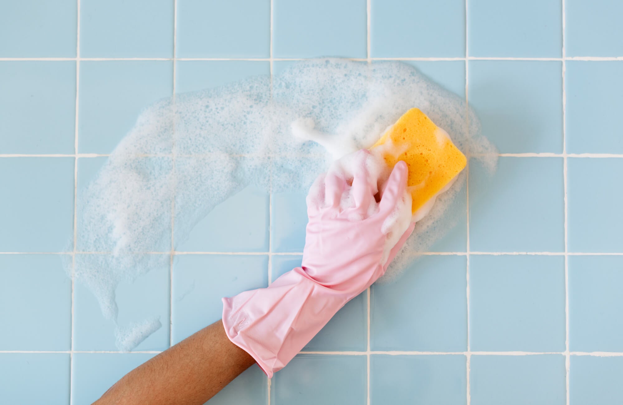 Close-up of a hand in a pink glove scrubbing pale blue backsplash tiles with a sponge and soap suds.