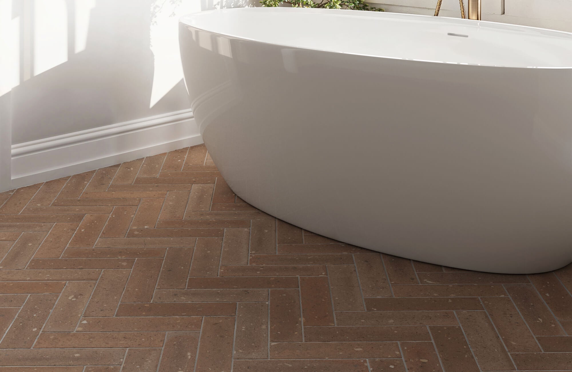 Matte terracotta look floor tiles in a herringbone pattern complement a sleek white freestanding bathtub in a modern bathroom.