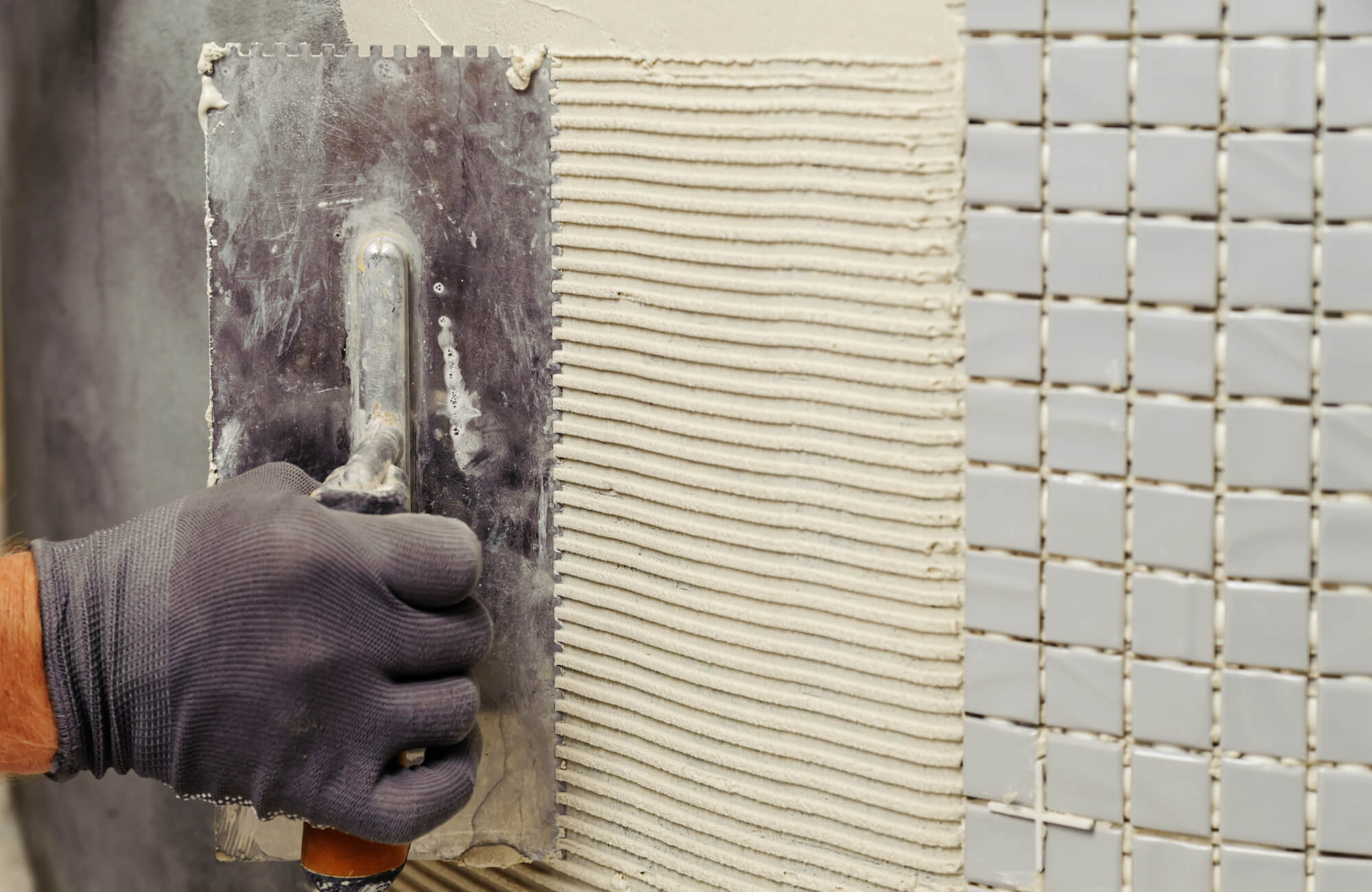 null Close-up of a gloved hand spreading thin-set mortar with a notched trowel while installing small square mosaic tiles on a wall surface.