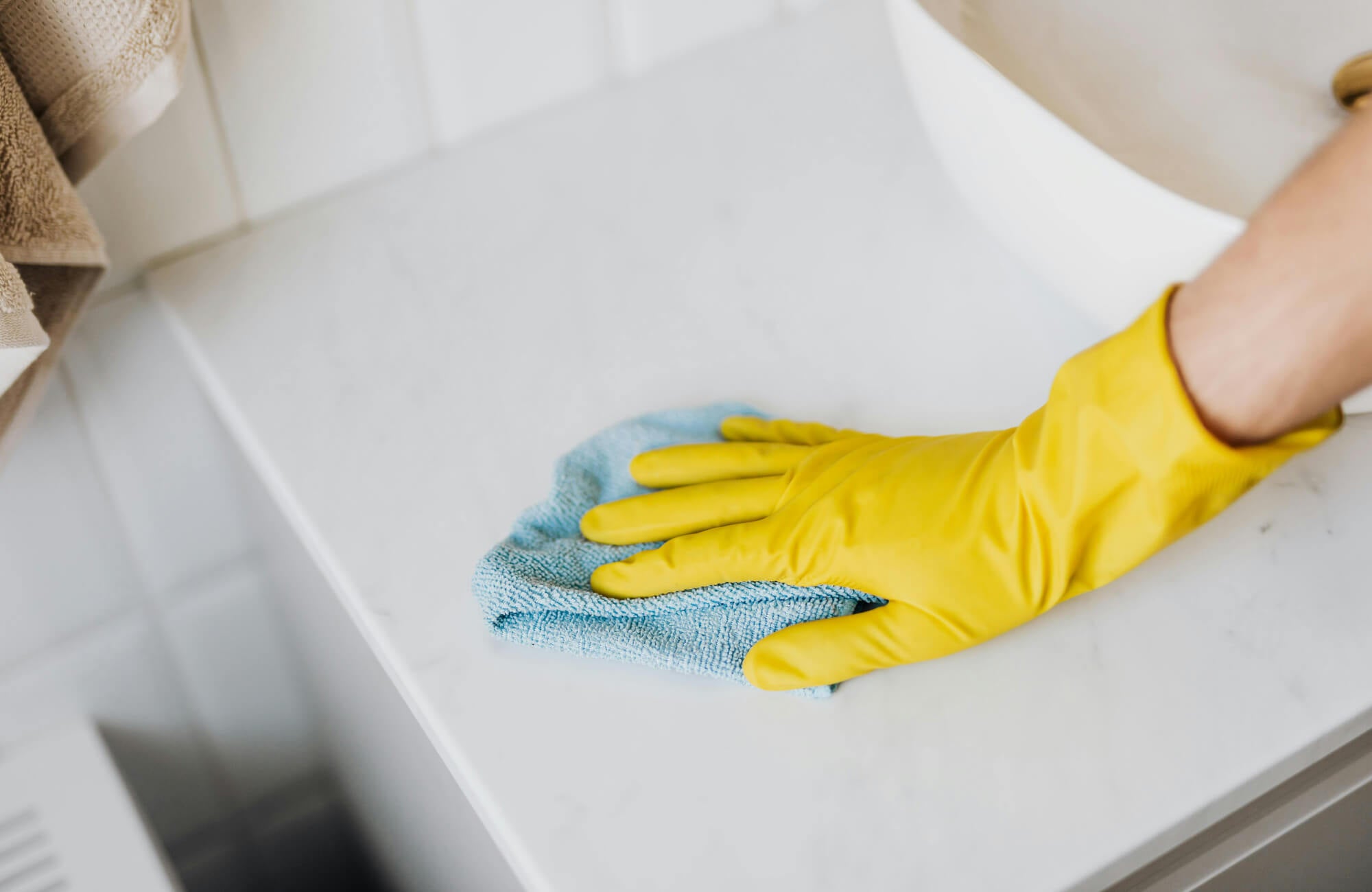 null Close-up of a hand in a yellow glove cleaning a white bathroom vanity countertop with a blue microfiber cloth.