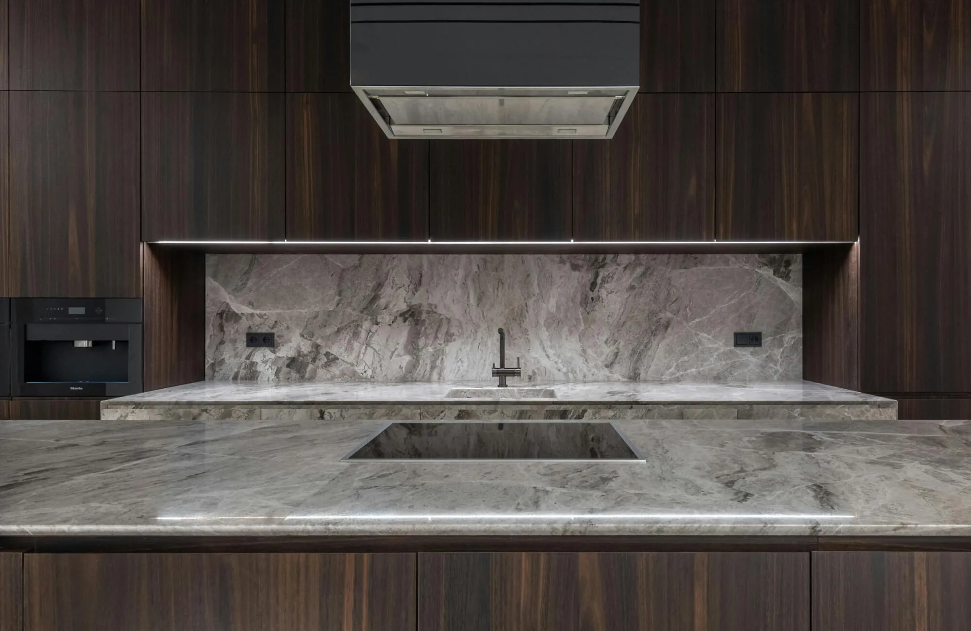 Full-height stone backsplash with dramatic gray veining adds depth and continuity behind a modern cooktop and sink area.